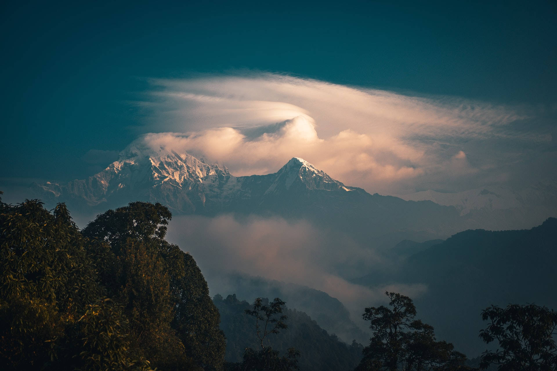 Mountain Landscape 桌布 壁紙 — 雄偉的雪山峰在黎明時分穿透發光的雲層，前景是黑暗的森林剪影。
