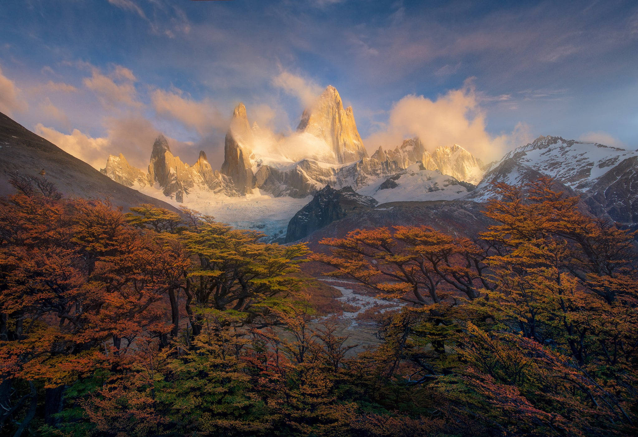 Patagonia 桌布 壁紙 — 雄偉的雪山峰穿透雲層，金色光芒沐浴著前景的秋樹。