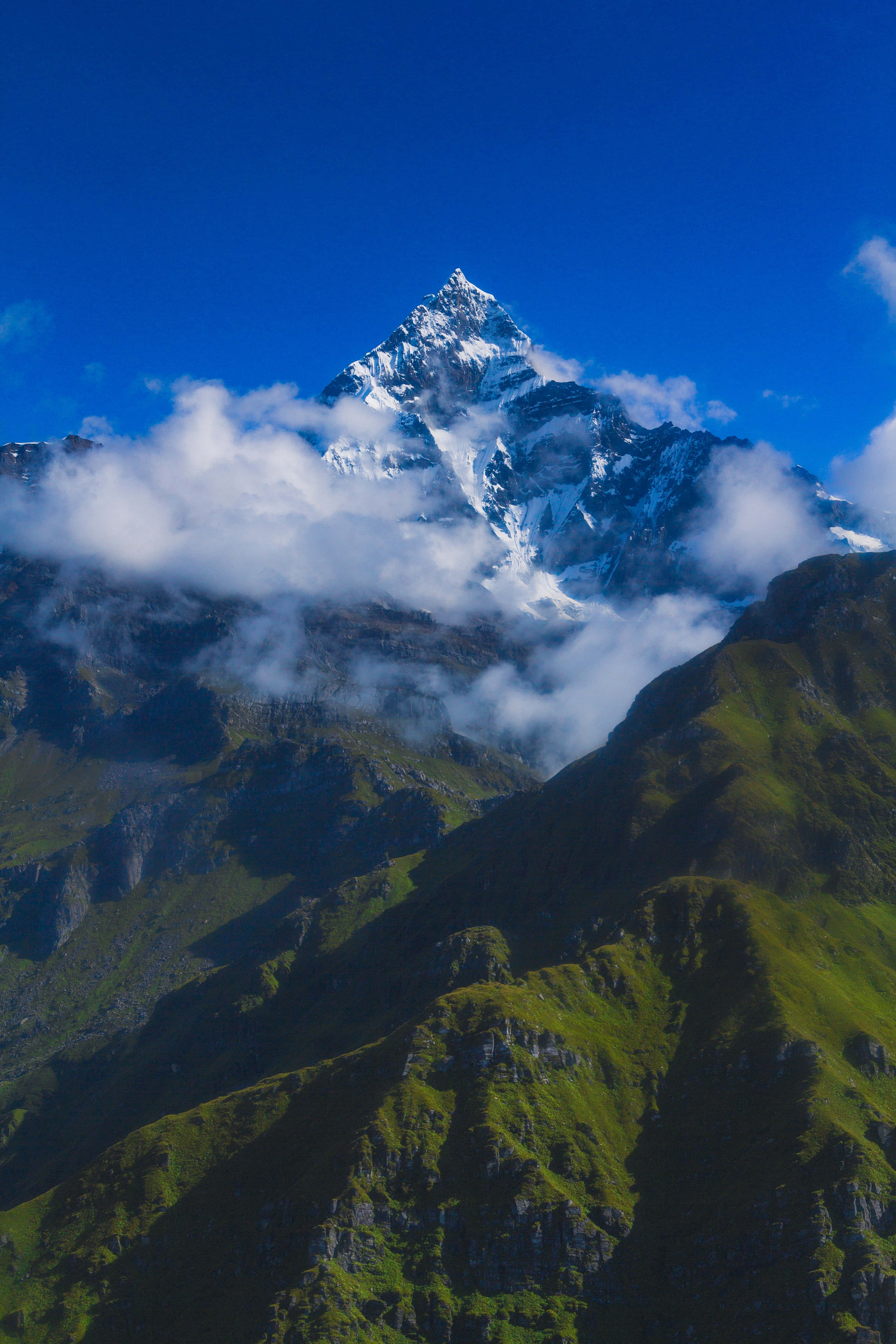 photo 桌布 壁紙 — 雄偉的雪山峰穿過蓬鬆的白雲，映襯著充滿活力的藍天，下方是鬱鬱蔥蔥的綠色山坡。