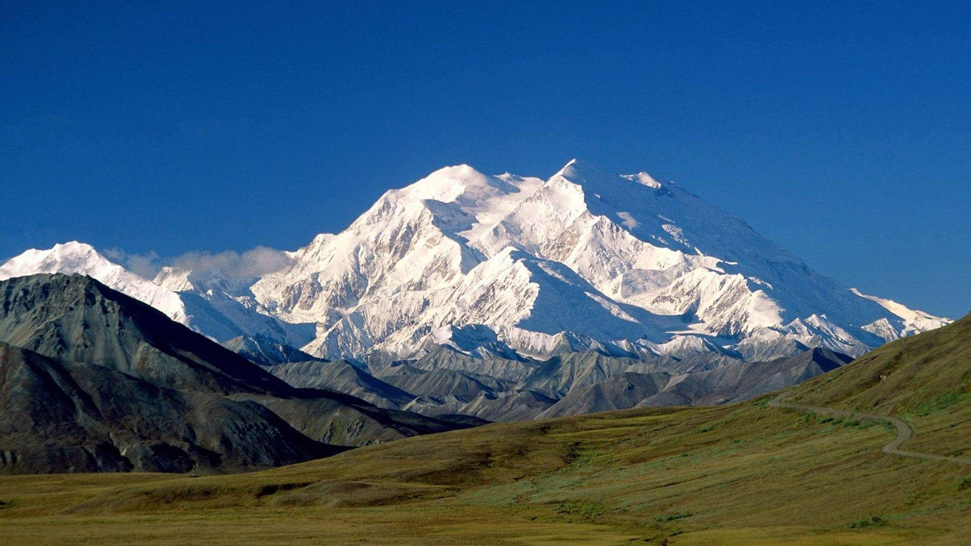 山脈 桌布 壁紙 — 雄偉的雪山峰頂在清澈的藍天下陡峭聳立，下方是崎嶇的山坡和蜿蜒的道路。