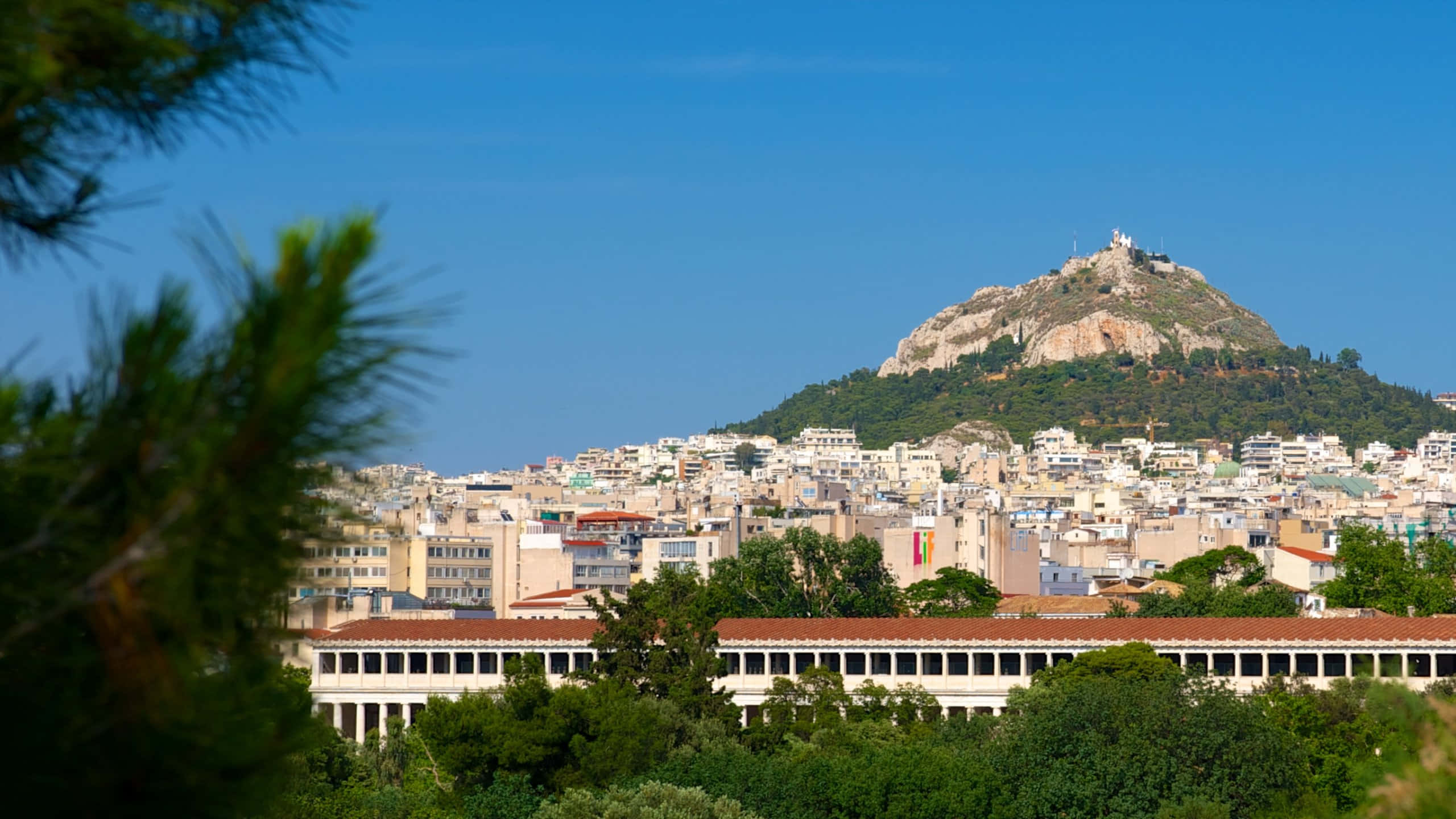 Blue Sky 桌布 壁紙 — 雅典充滿活力的城市景觀，衛城山丘在密集建築物上方聳立，天空湛藍清澈。