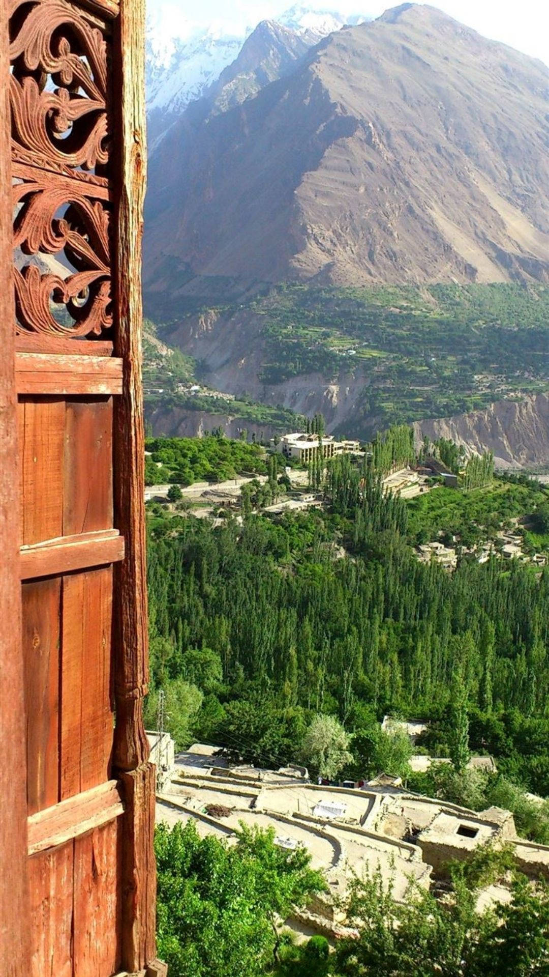 Pakistan 桌布 壁紙 — 雕刻木窗框住了一個令人嘆為觀止的景色，一個翠綠山谷坐落於高聳的雪山之間。