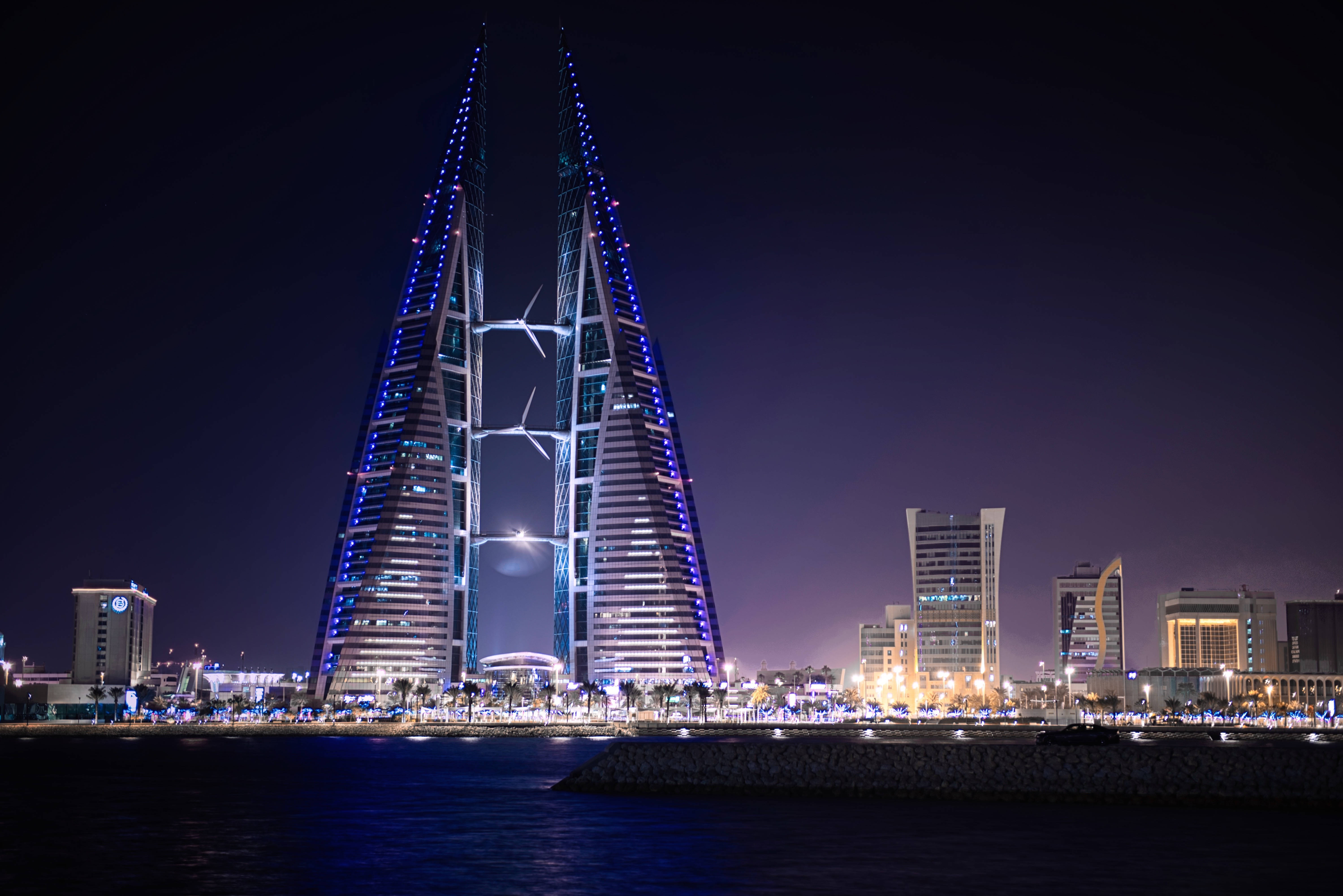 Bahrain 桌布 壁紙 — 雙子摩天大樓在黑暗的夜空中發出藍光，映照在海濱城市景觀之上。