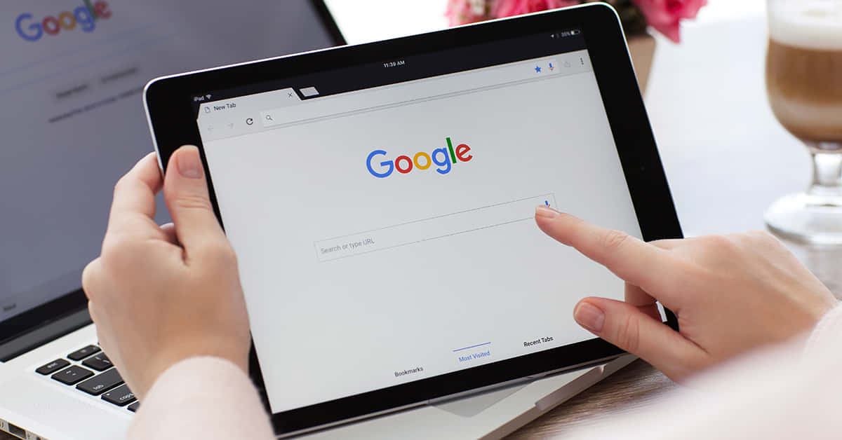 有人在附近有筆記型電腦和咖啡的情況下使用平板電腦搜尋Google。 — Google Meet 壁紙