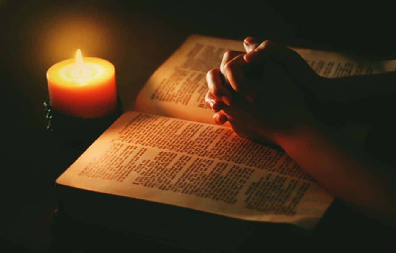 Praying 桌布 壁紙 — 雙手合十祈禱，放在一本打開的書上，旁邊有一根在昏暗光線中燃燒的蠟燭。