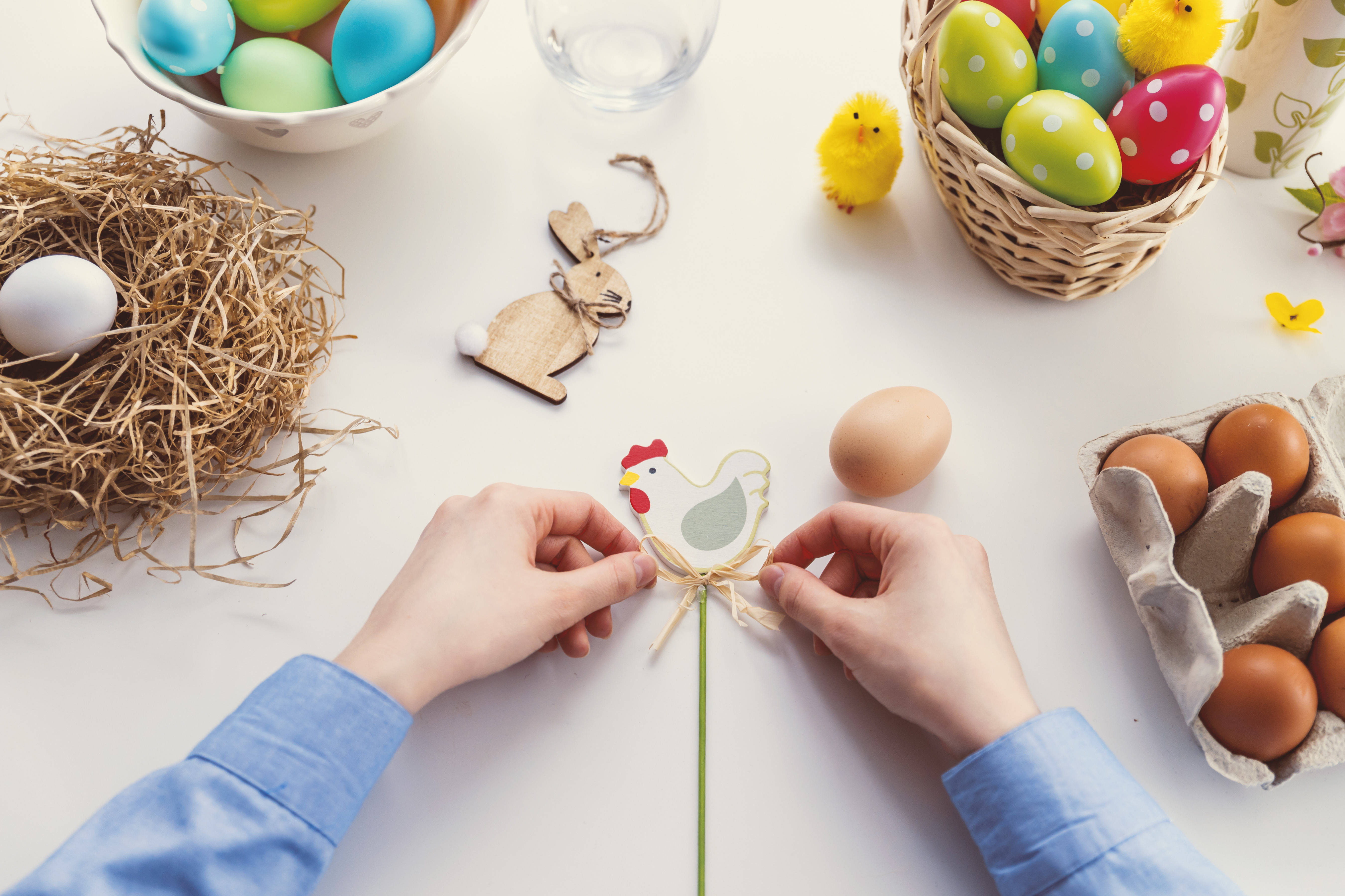 Happy Easter 桌布 壁紙 — 雙手在明亮的白色桌面上，於色彩繽紛的復活節蛋、巢穴和小雞之間組裝紙雞裝飾。