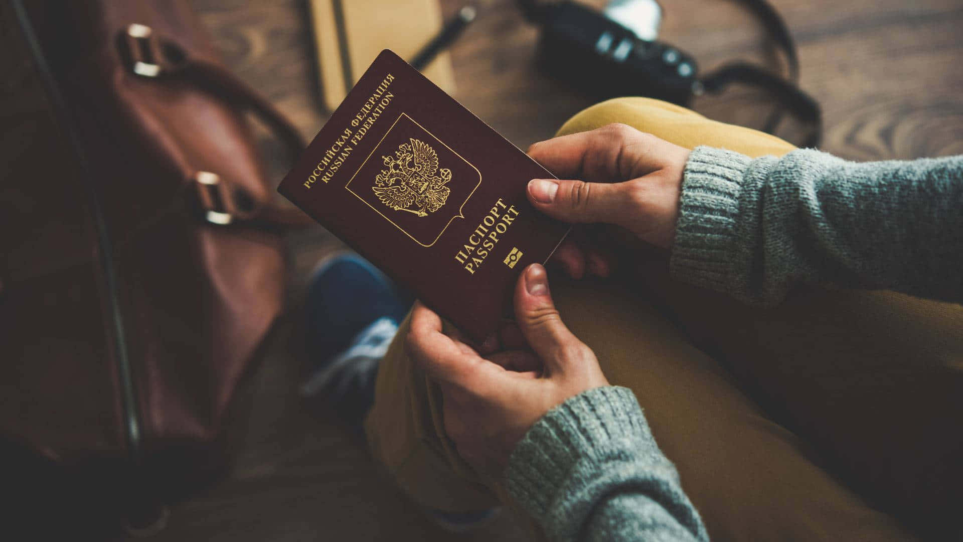 Passport 桌布 壁紙 — 雙手在木地板上方，旁邊有一個皮袋和相機，手持俄羅斯護照。