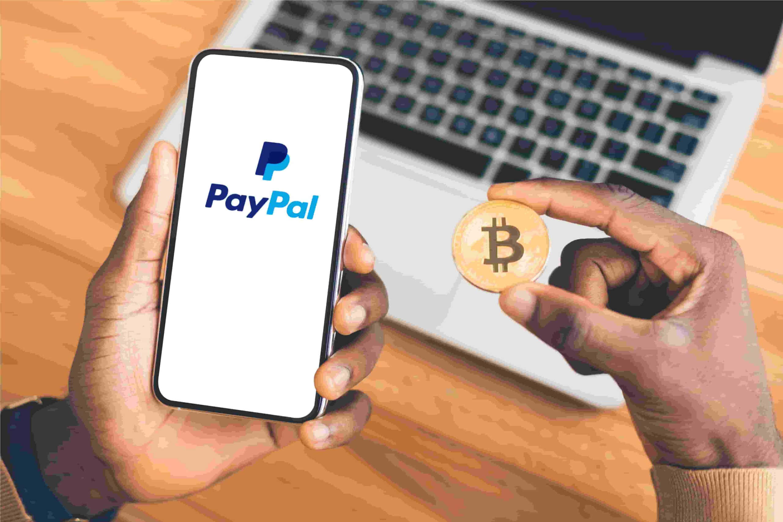 PayPal 桌布 壁紙 — 雙手在木桌上拿著一部顯示PayPal標誌的智慧型手機和一枚實體比特幣硬幣，放在筆記型電腦上方。