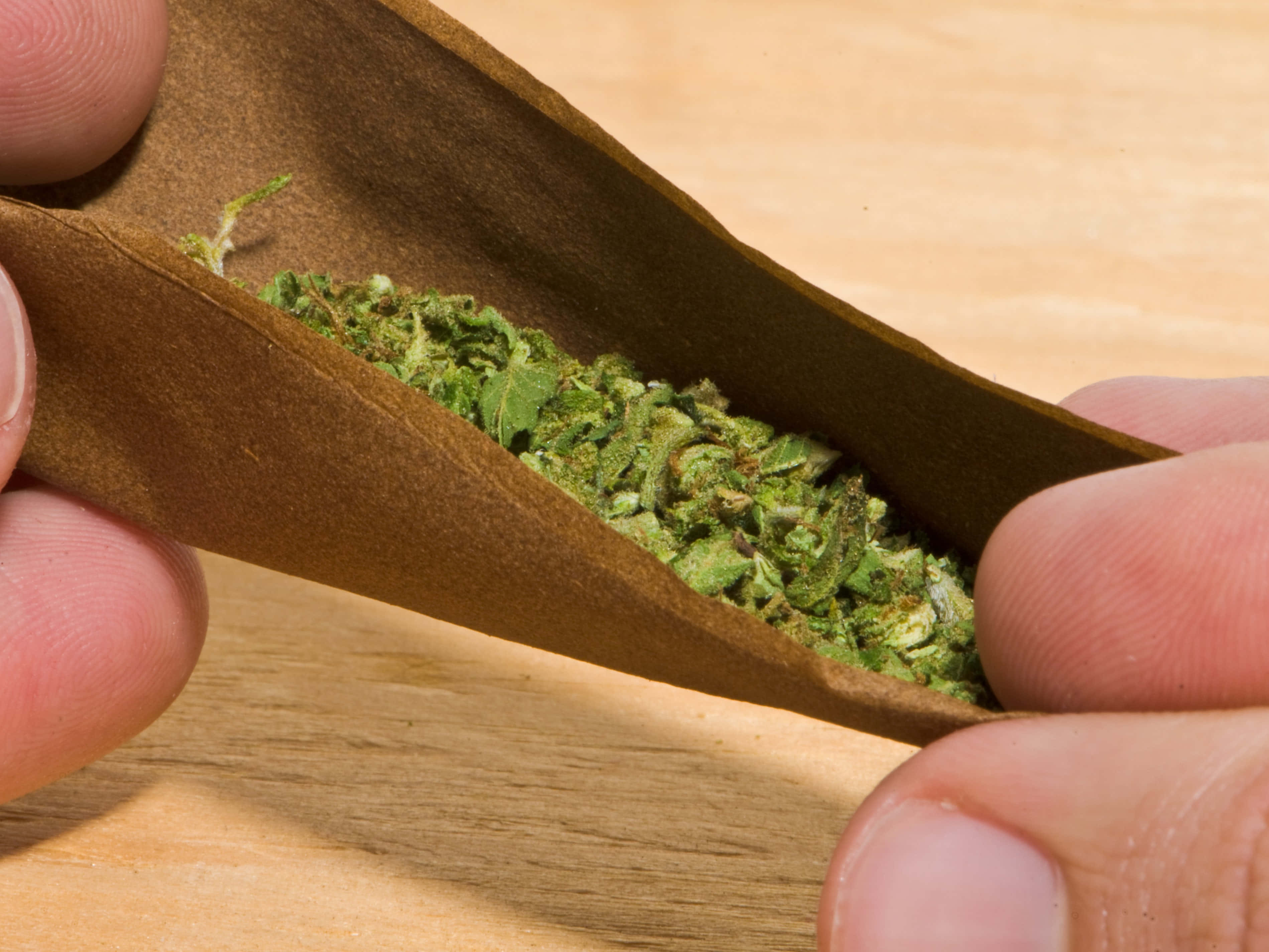 Weed Blunt 桌布 壁紙 — 雙手在木質表面上捲起裝滿綠色大麻花蕾的棕色紙煙的特寫。