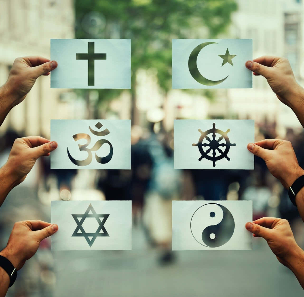 Theology 桌布 壁紙 — 雙手在模糊的城市街道背景下，手持六個宗教符號，包括十字架、新月、星星、嗡、法輪和陰陽。