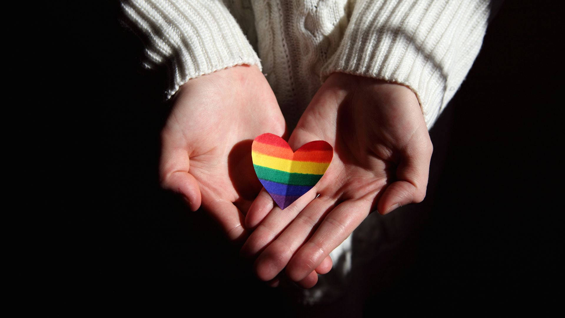 Subtle Lgbt 桌布 壁紙 — 雙手在深色背景前捧著彩虹心，柔和光線突顯這個姿勢。