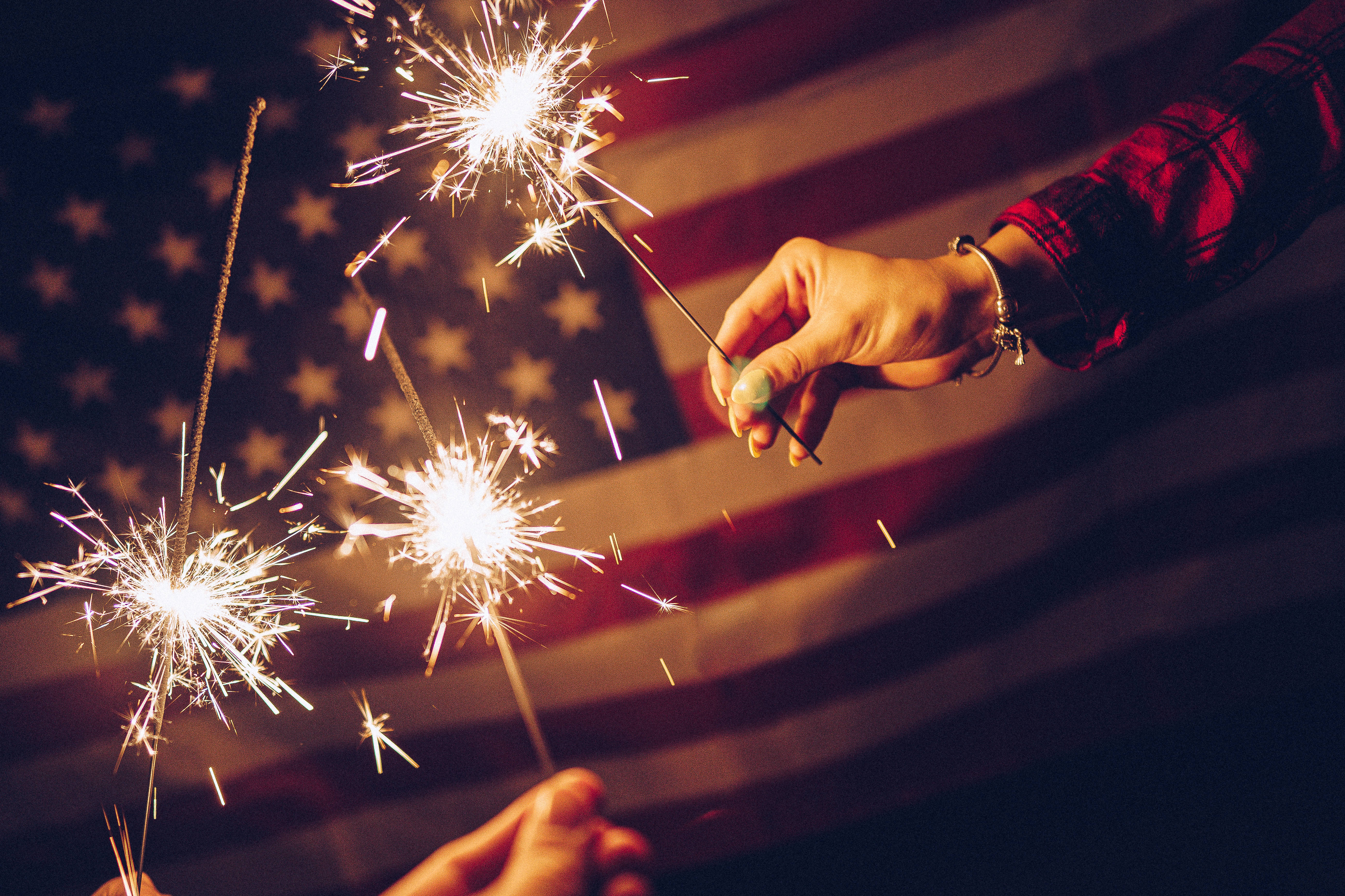 USA flag iPhone 桌布 壁紙 — 雙手在溫暖的夜間燈光下，手持閃爍的煙火，背景是模糊的美國國旗。