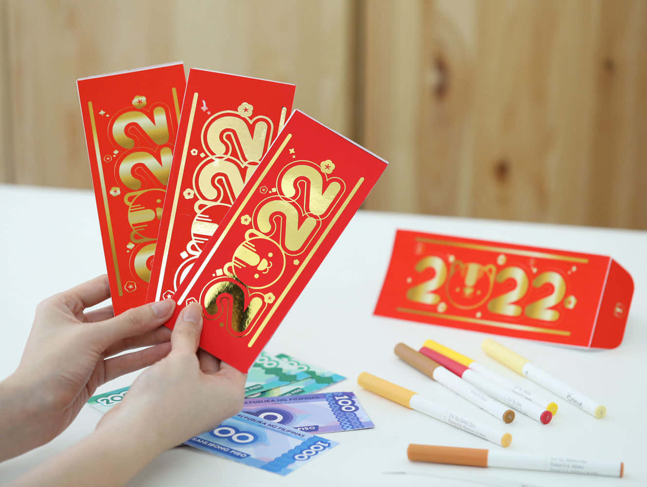 Red Envelope 桌布 壁紙 — 雙手在白色桌子上，拿著帶有金色2022設計的紅色紅包，放在貨幣和記號筆上方。