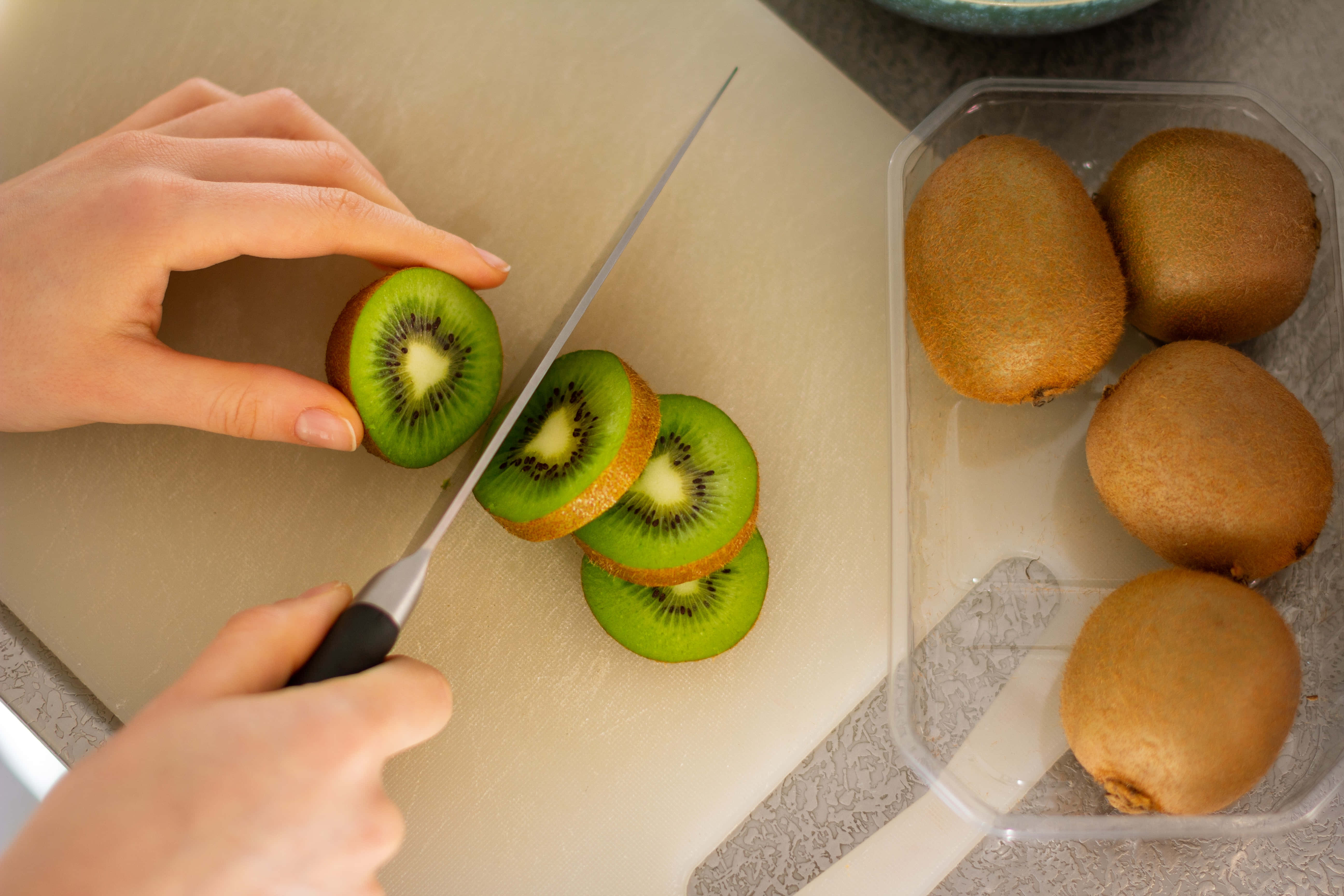 Kiwi 桌布 壁紙 — 雙手在米色砧板上切新鮮奇異果的特寫，附近塑膠容器內有完整奇異果。