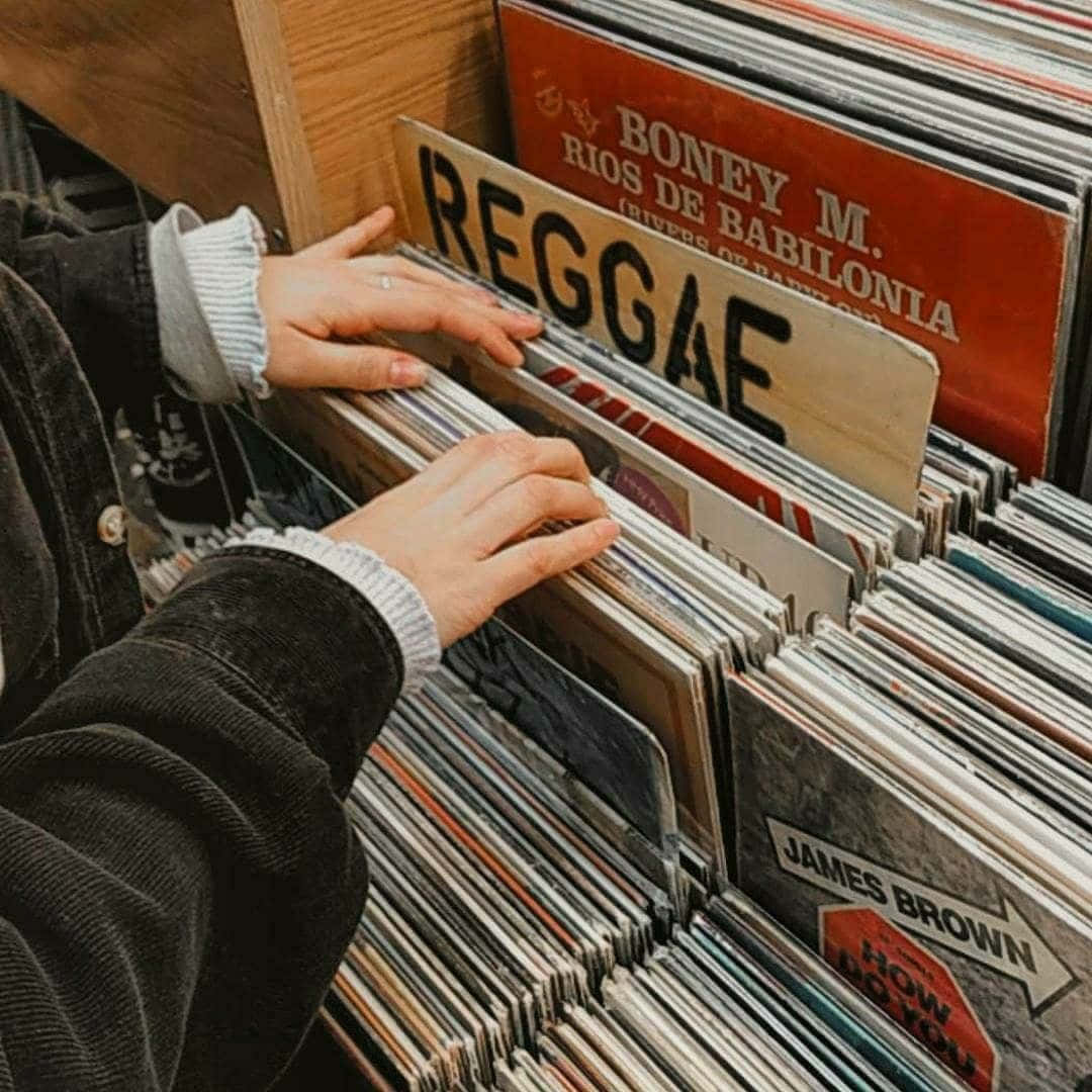 復古美學 桌布 壁紙 — 雙手在舒適的唱片店裡，在 'REGGAE' 標誌下翻閱復古黑膠唱片，店內充滿經典音樂。
