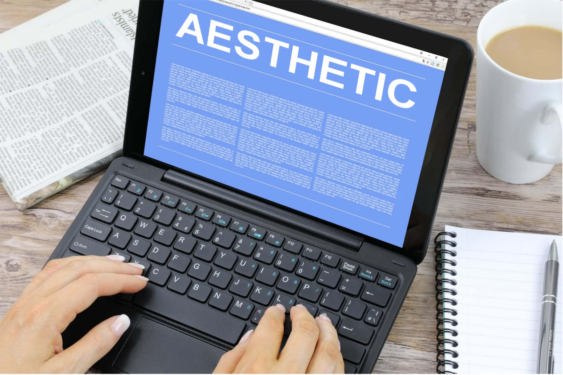 美學筆電 桌布 壁紙 — 雙手在顯示「aesthetic」一詞的筆記型電腦上打字，旁邊的木桌上放著一杯咖啡和一本筆記本。