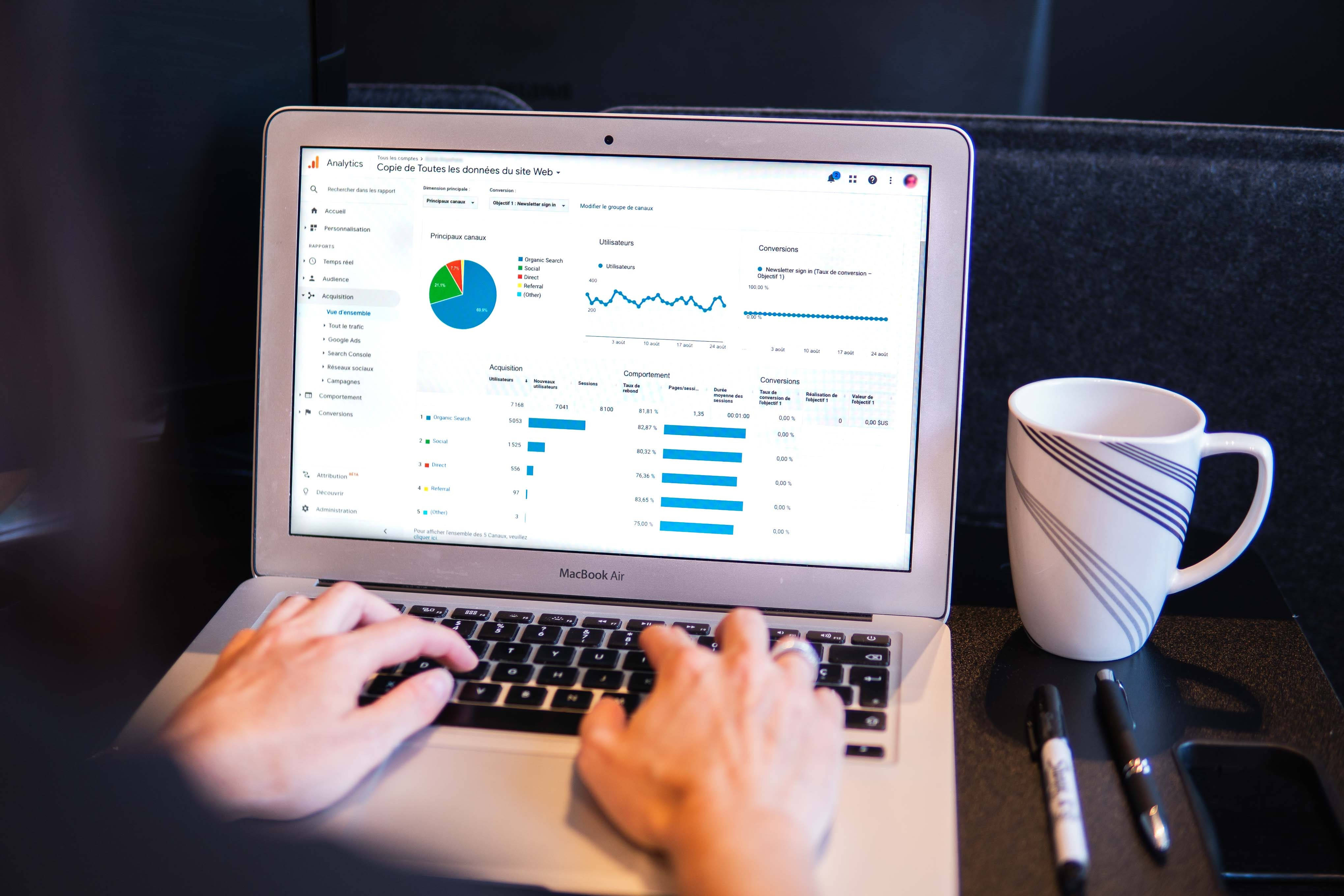 Sales 桌布 壁紙 — 雙手在顯示分析儀表板的筆記型電腦上打字，旁邊有一個條紋馬克杯在深色桌子上。