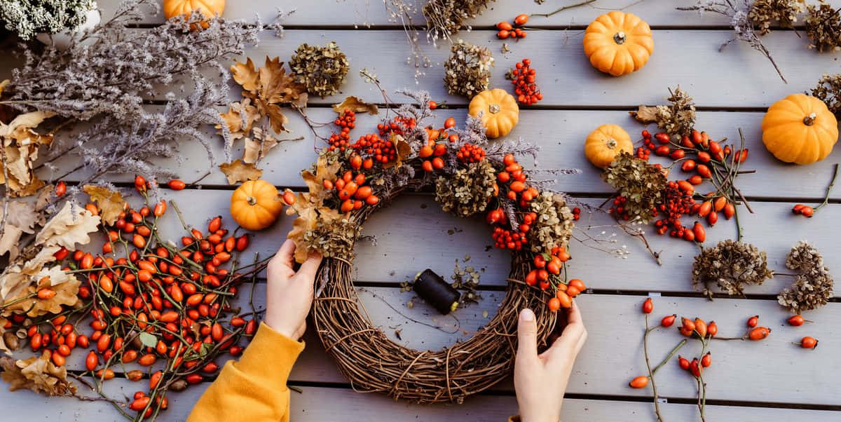 Fall Thanksgiving 桌布 壁紙 — 雙手在風化的木質表面上製作一個質樸的秋季花環，飾有橙色漿果、南瓜和乾燥樹葉。