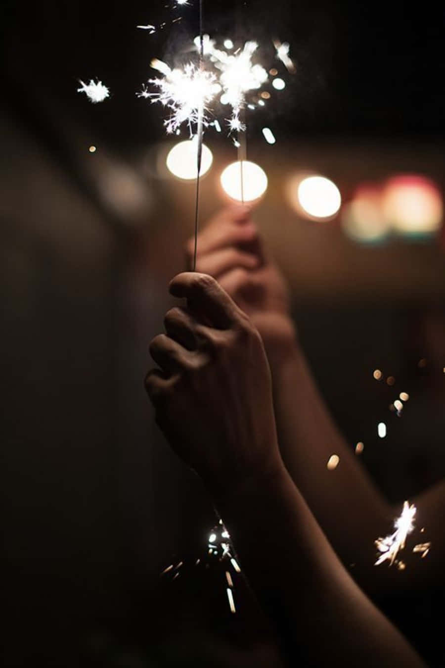 Celebration 桌布 壁紙 — 雙手在黑暗背景前拿著閃爍的煙火，背後有柔和發光的燈光。