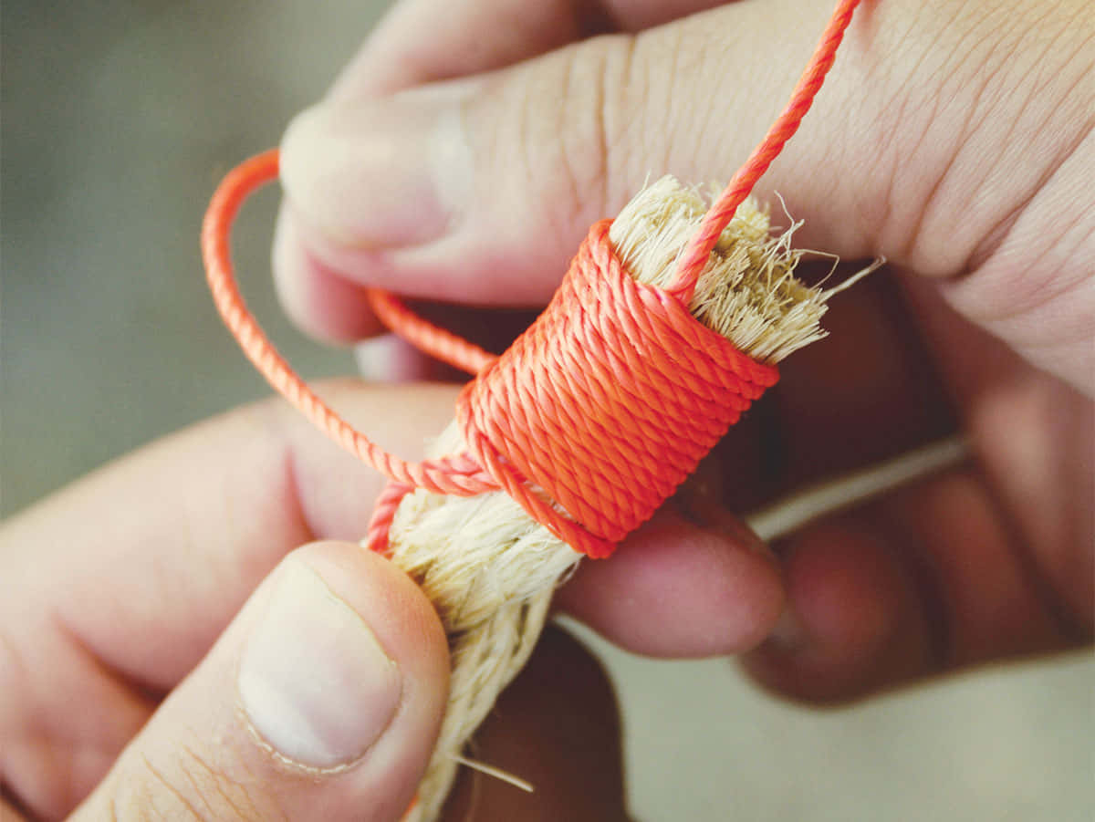 whip 桌布 壁紙 — 雙手將亮橙色繩索精準而專注地纏繞在一束天然纖維上的特寫。