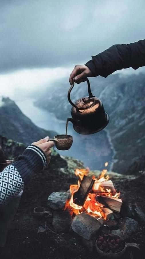 Winter Camping 桌布 壁紙 — 雙手將水壺中的熱茶倒在噼啪作響的營火上，背景是霧氣繚繞的山巒。