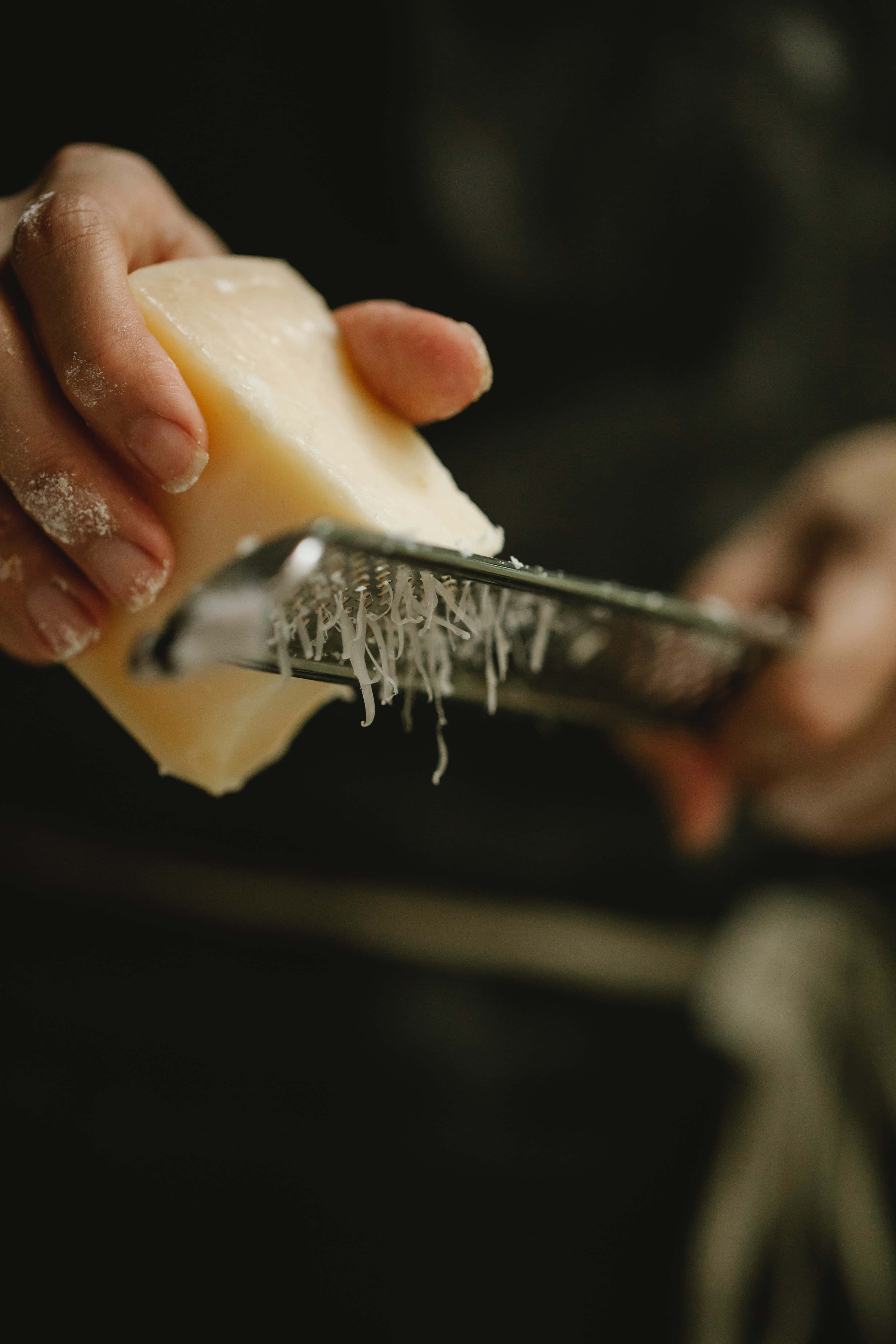 Cheese 桌布 壁紙 — 雙手將淡色起司塊在金屬刨絲器上刨絲的特寫，細碎的起司絲落下。