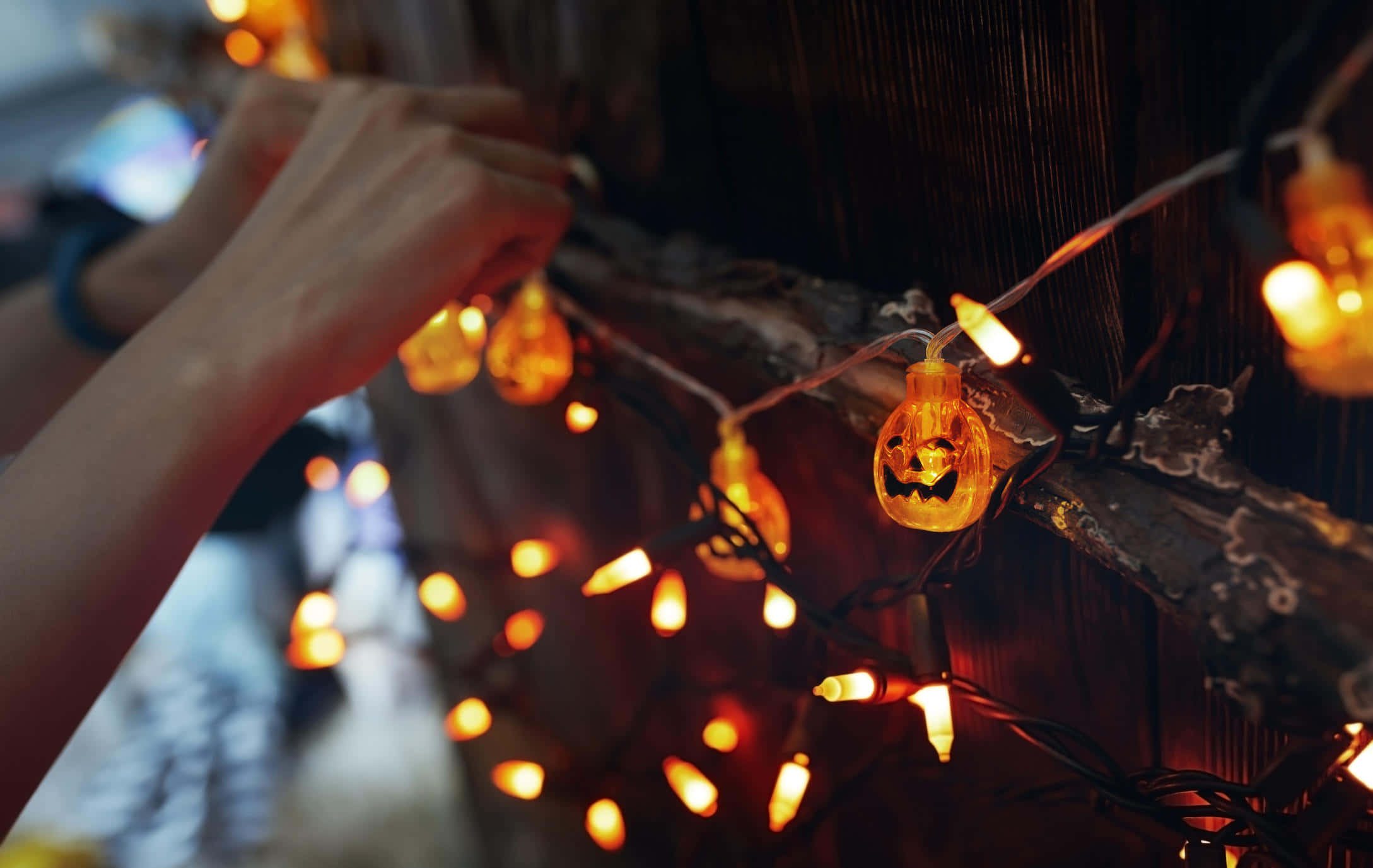 Halloween Yard Decorations 桌布 壁紙 — 雙手將發光的傑克南瓜燈串燈掛在溫暖琥珀色調的鄉村木樑上。