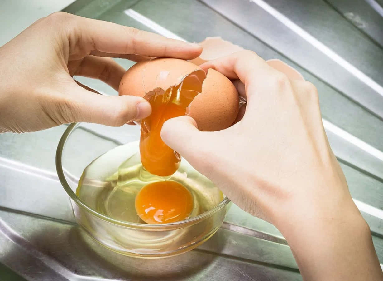 Egg 桌布 壁紙 — 雙手小心地將棕色雞蛋敲入玻璃碗中，在不鏽鋼表面上露出鮮豔的橙色蛋黃和清澈的蛋白。
