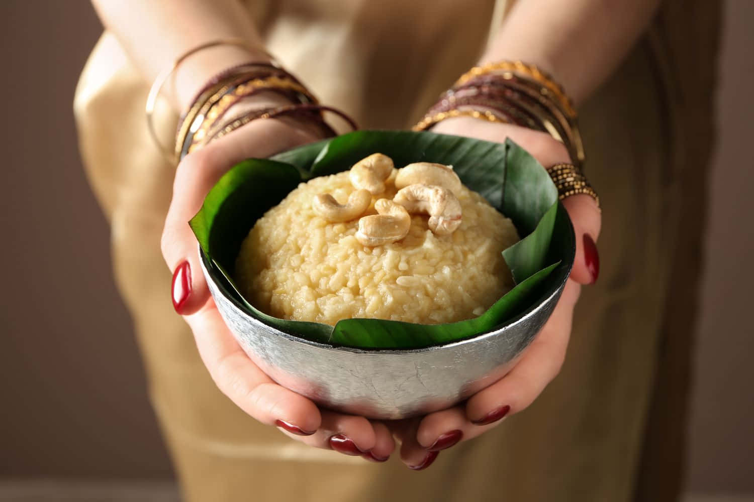 Pongal 桌布 壁紙 — 雙手戴著手鐲和紅色指甲油，輕輕捧著一個銀碗，裡面盛著蒸米飯，上面放著腰果，放在一片綠葉上。