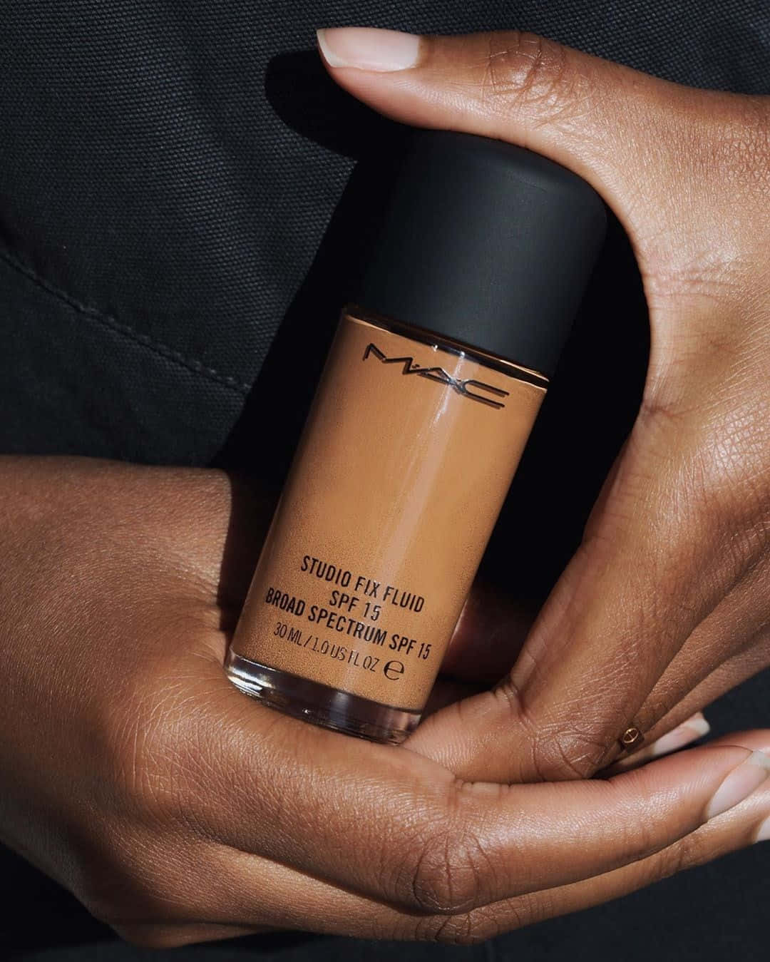 Mac Cosmetics 桌布 壁紙 — 雙手拿著MAC Studio Fix Fluid粉底液瓶子的特寫，背景為深色紋理。