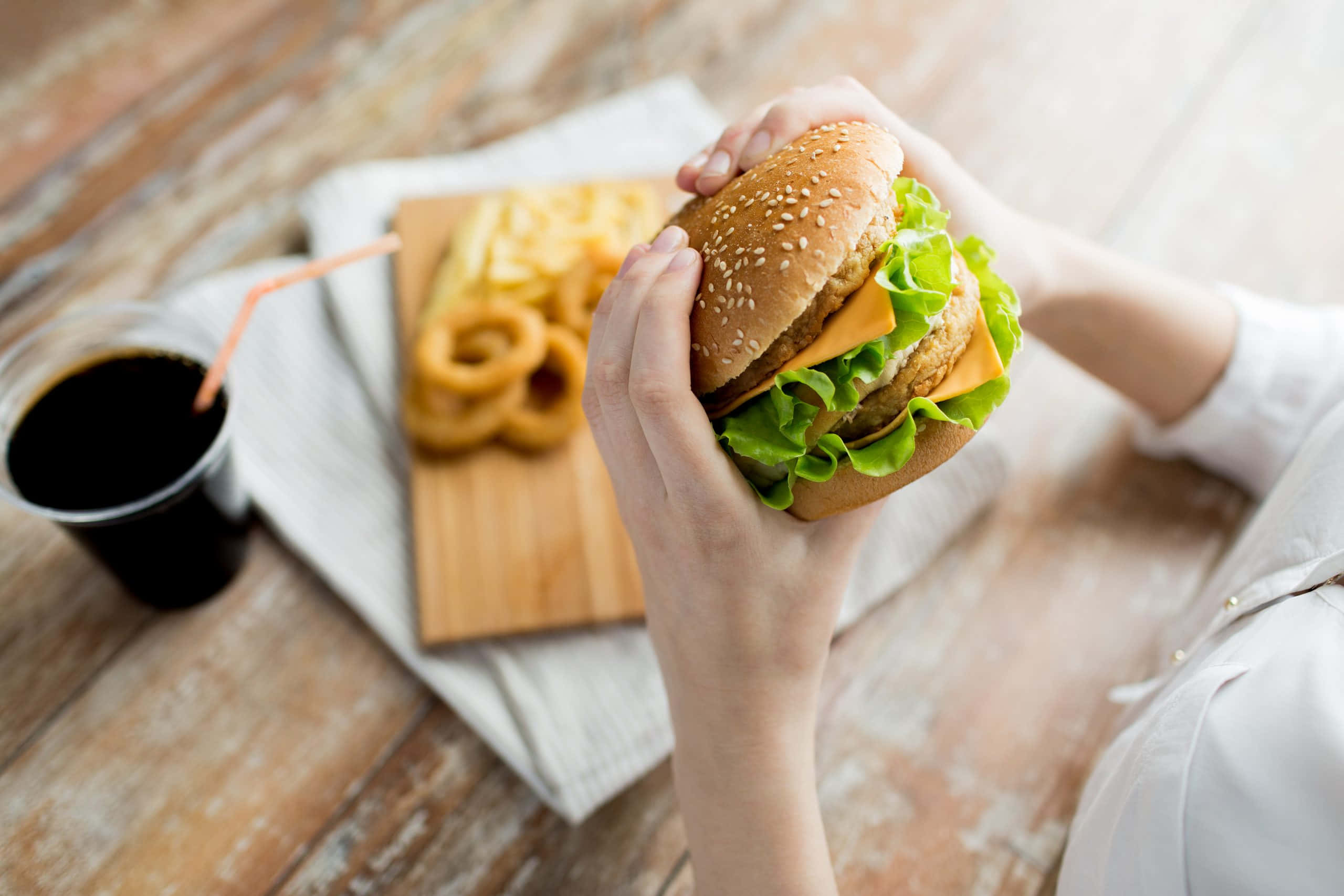 fast food 桌布 壁紙 — 雙手拿著一個多汁的芝麻漢堡，裡面有生菜、起司和肉餅，放在木桌上，旁邊有薯條、洋蔥圈和汽水。