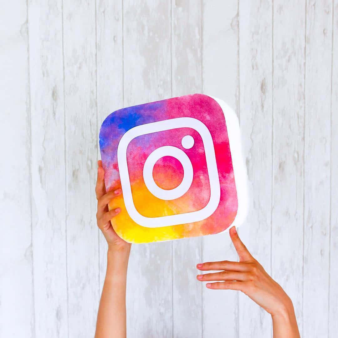 Logo Design 桌布 壁紙 — 雙手拿著一個色彩繽紛的水彩Instagram標誌毛絨玩具，背景是白色木牆。