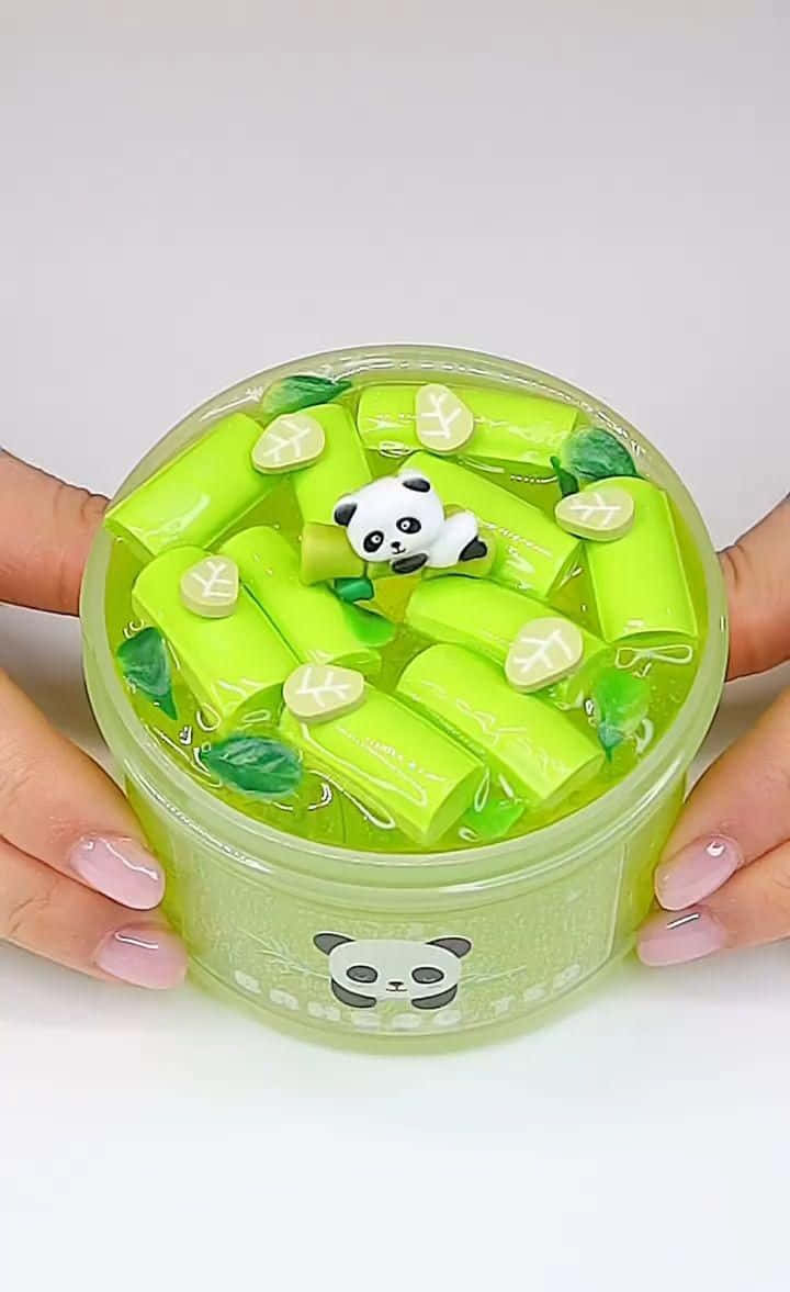 熊貓 桌布 壁紙 — 雙手拿著一個透明容器，裡面裝滿了形狀像竹莖和竹葉的亮綠色黏液，還有一個小熊貓公仔漂浮在裡面。