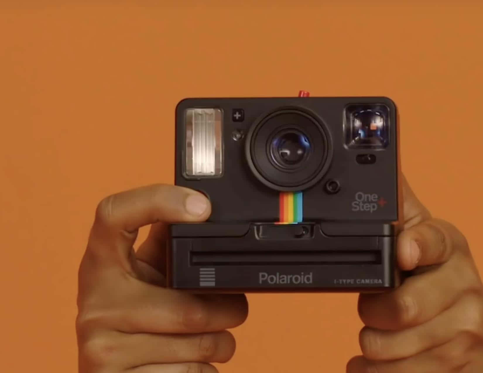 Photograph 桌布 壁紙 — 雙手拿著一台黑色Polaroid One Step+拍立得相機，背景為純橙色。
