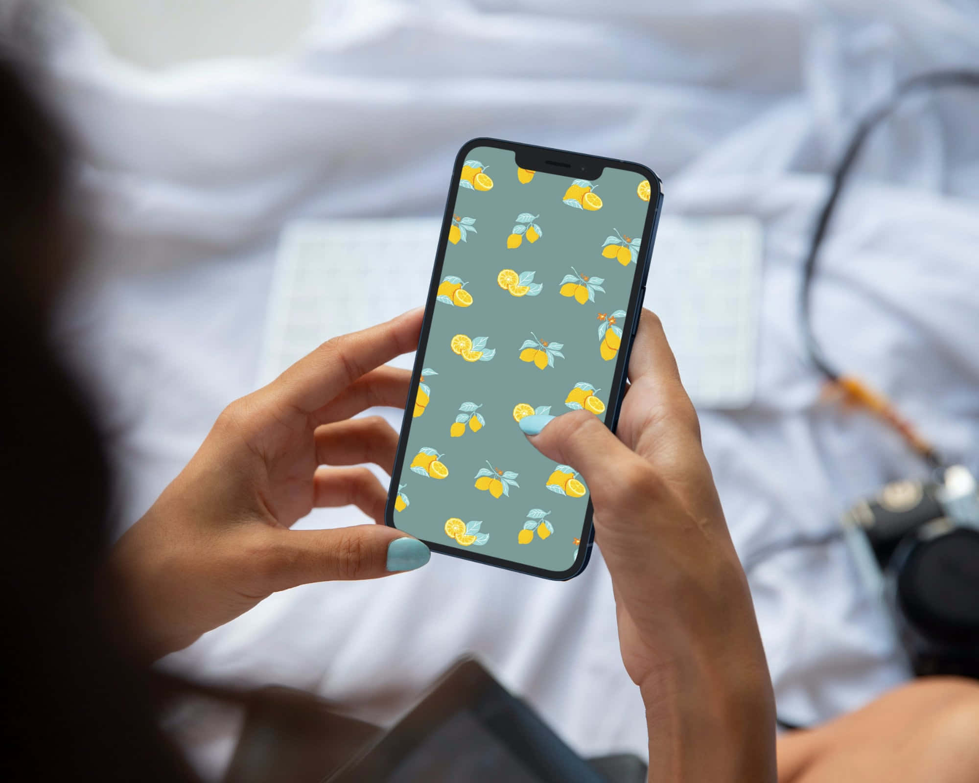 Lemon iPhone 桌布 壁紙 — 雙手拿著一部手機，在柔軟的白色床罩上顯示檸檬圖案壁紙。