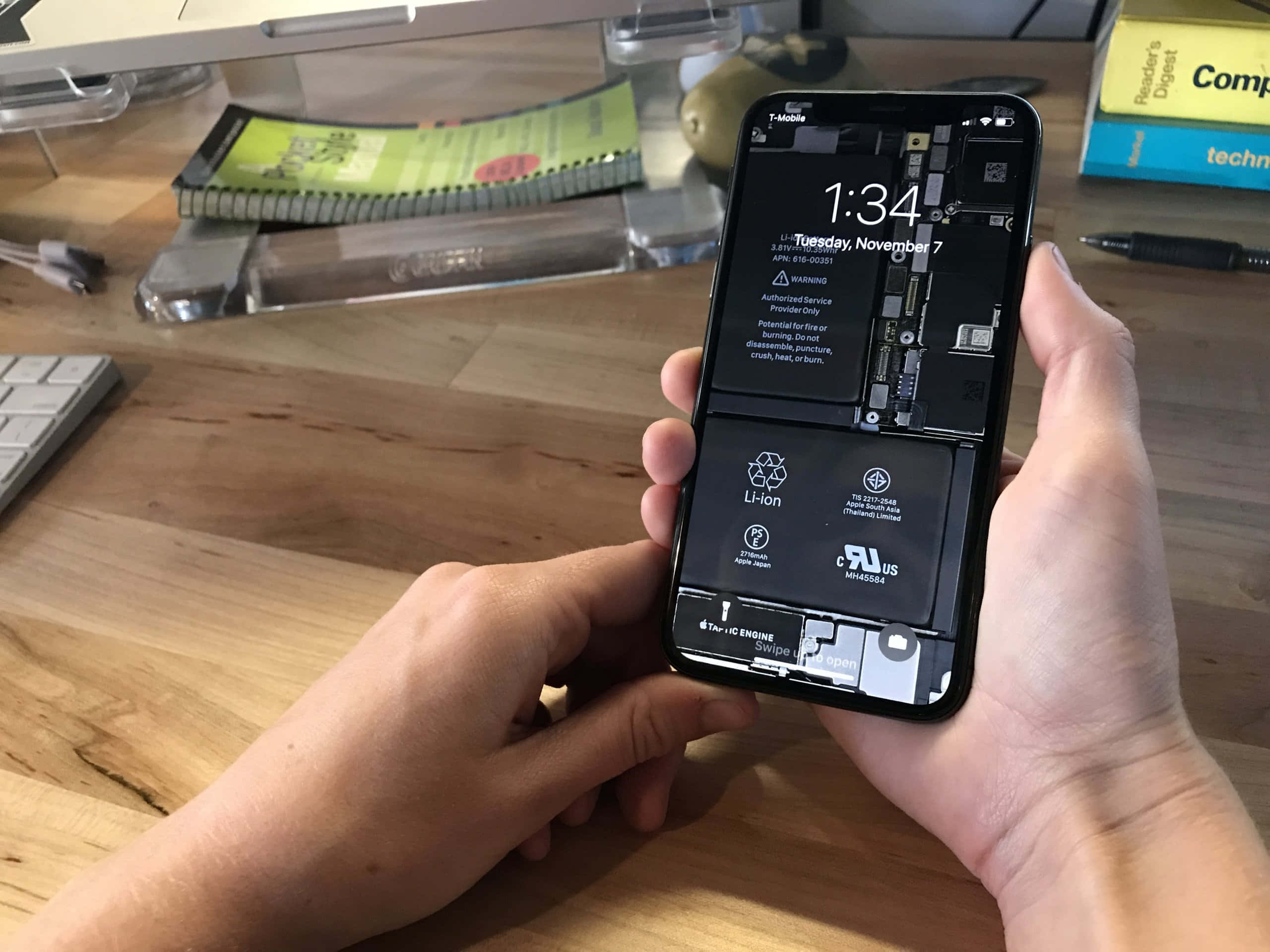 手機 桌布 壁紙 — 雙手拿著一部拆開的iPhone，在木桌上露出內部零件和電池，旁邊有科技物品。