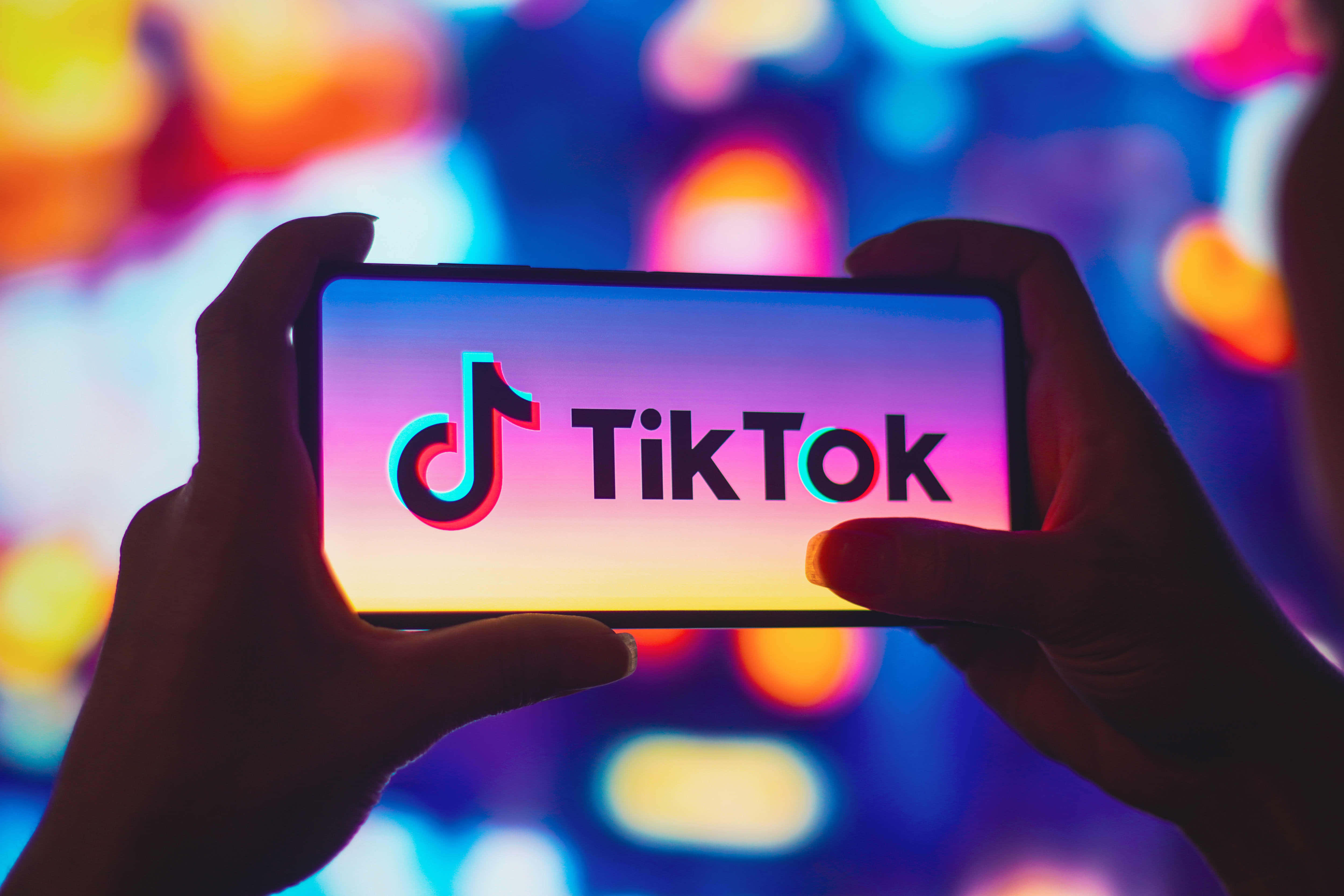 TikTok 動圖 桌布 壁紙 — 雙手拿著一部顯示TikTok標誌的智慧型手機，背景是色彩繽紛的燈光散景。