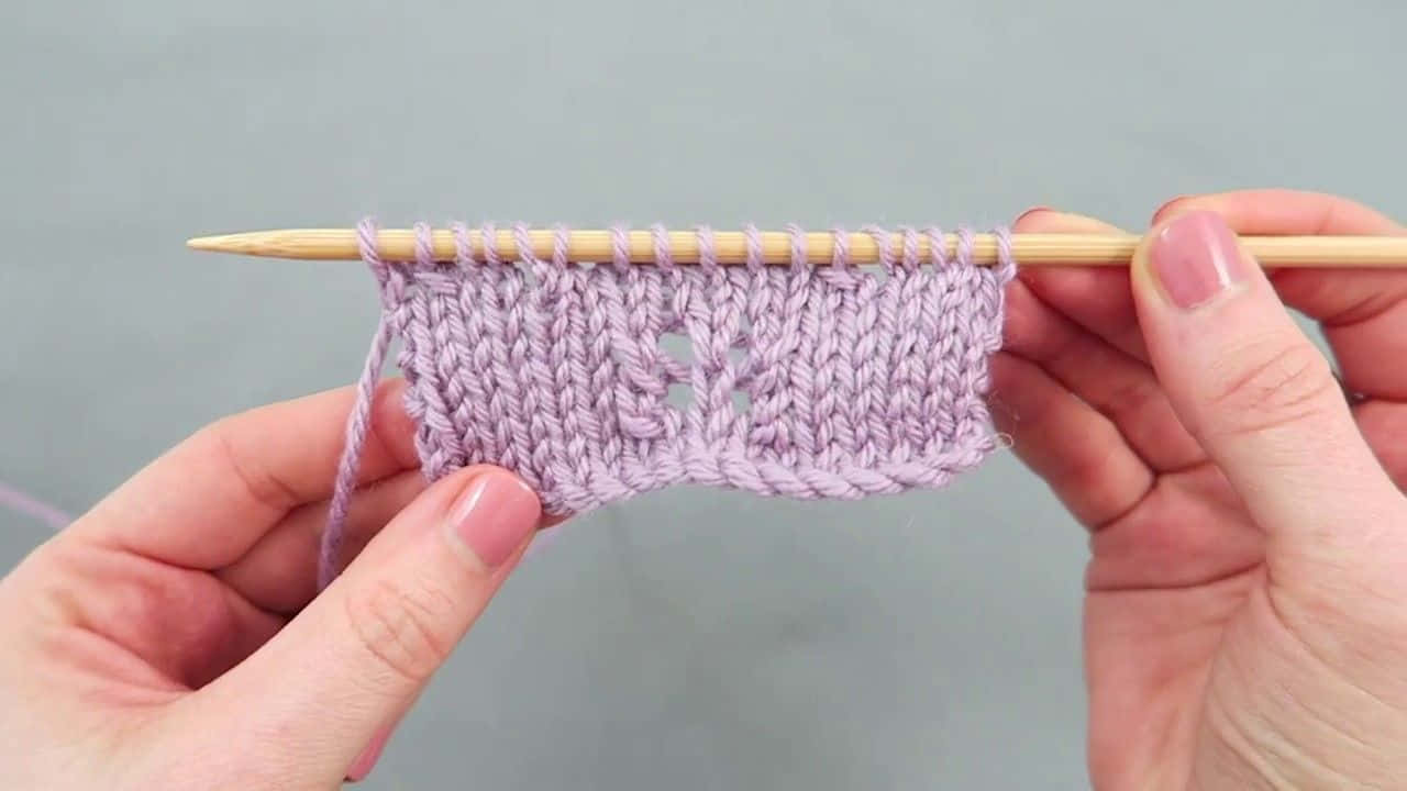 Butterfly Stitch 桌布 壁紙 — 雙手拿著木製編織針，上面有薰衣草色編織樣本，帶有蕾絲圖案，背景為灰色。