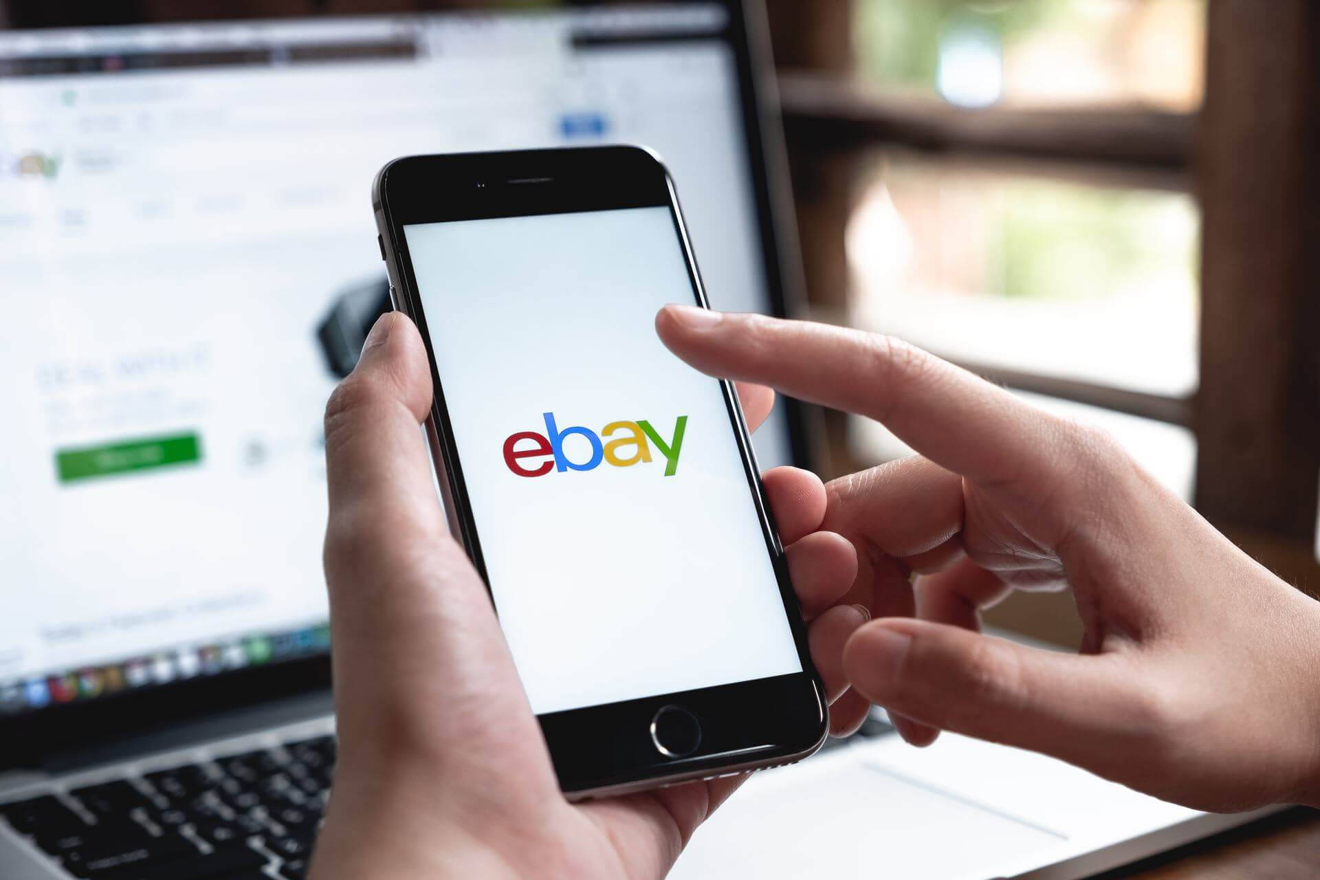 eBay 桌布 壁紙 — 雙手拿著顯示eBay標誌的智慧型手機，背景中可見同一網站的筆記型電腦。