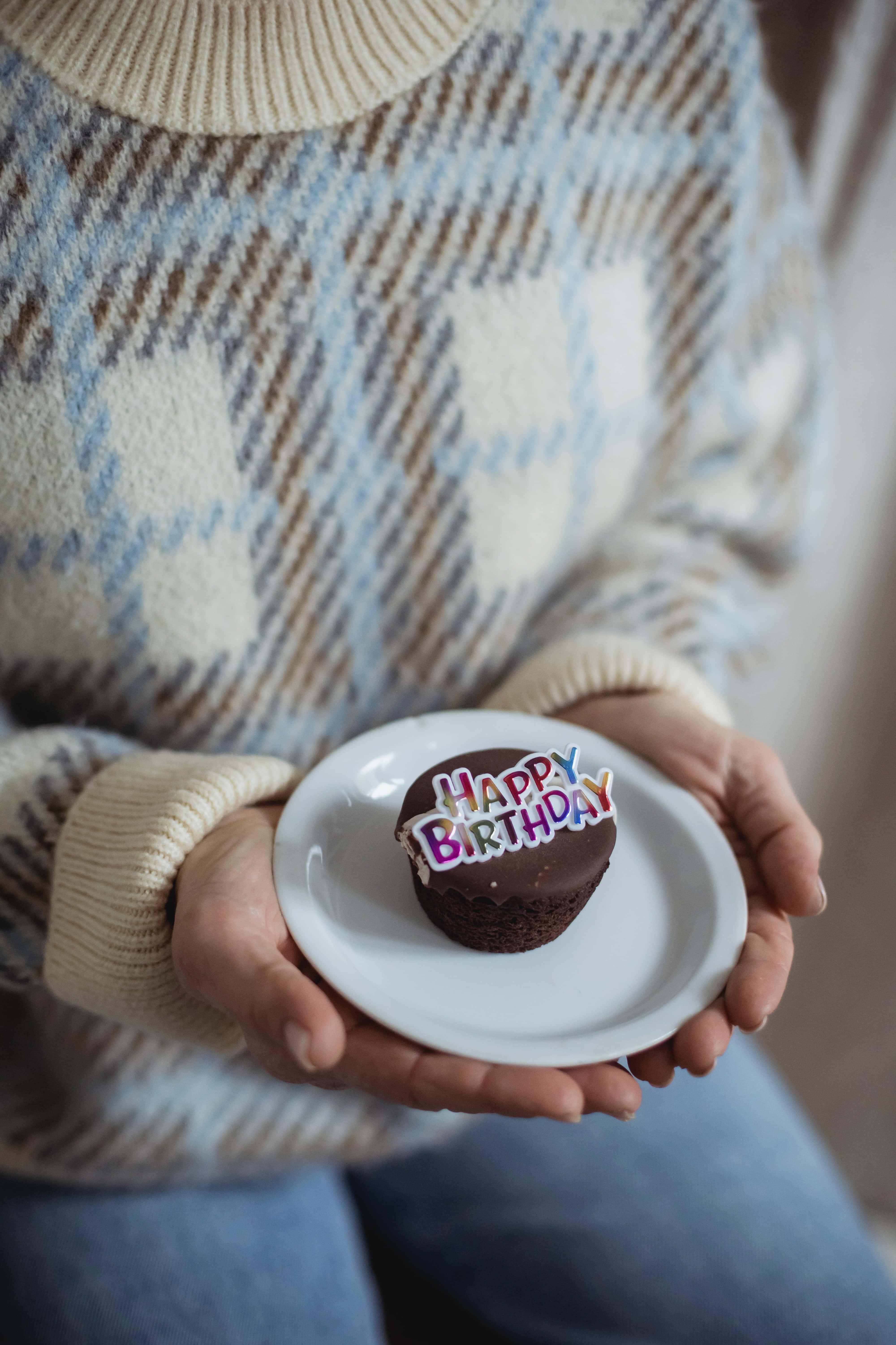 Cute Birthday 桌布 壁紙 — 雙手捧著一個白色盤子，上面放著一個巧克力杯子蛋糕，頂部綴有色彩繽紛的「Happy Birthday」糖霜。