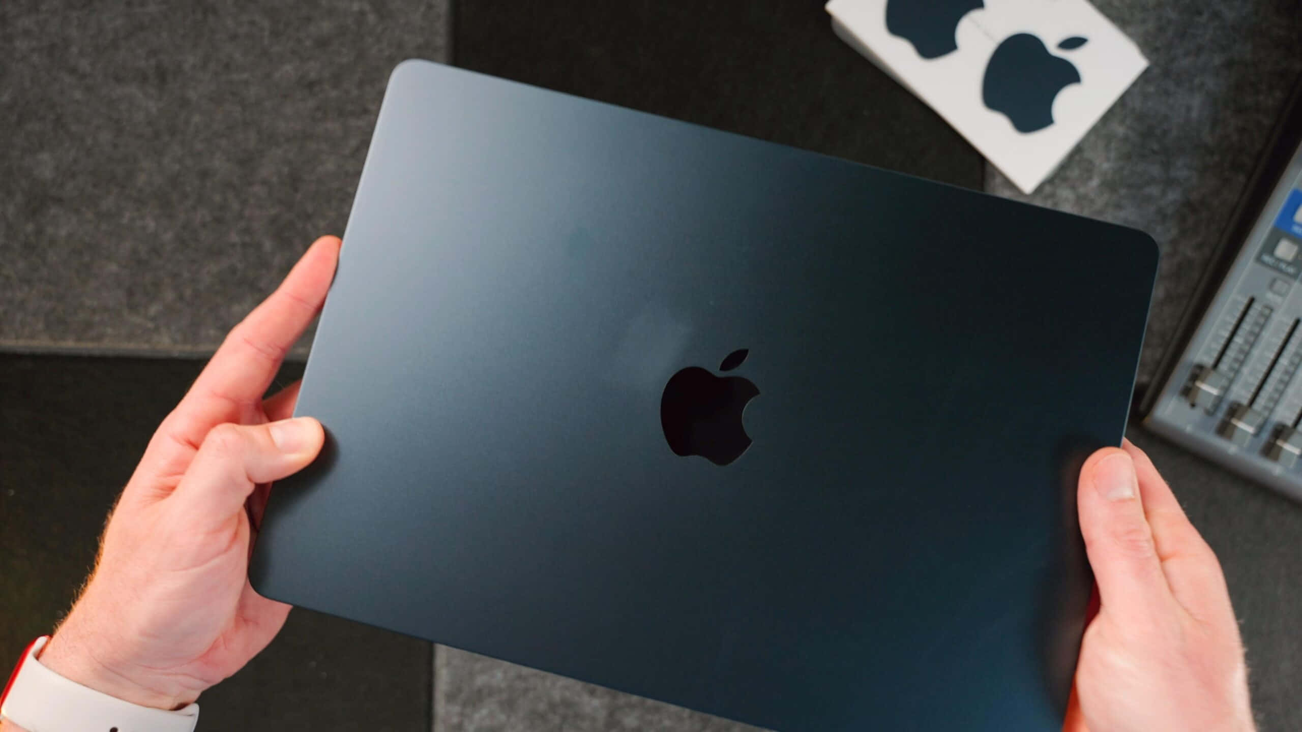 Black Macbook 桌布 壁紙 — 雙手捧著一台纖薄的霧面黑色 MacBook，Apple 標誌居中，背景是貼有貼紙和音響設備的深色紋理桌面。