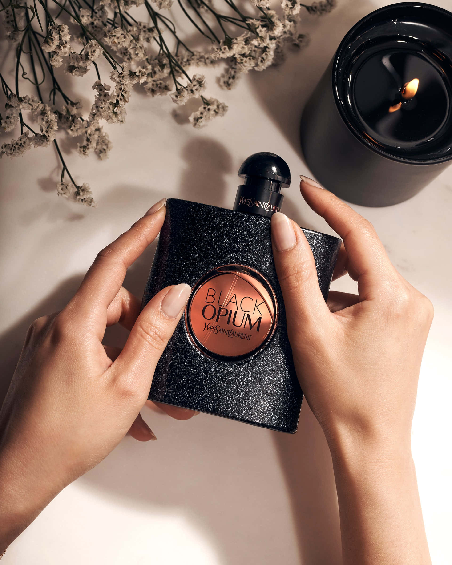 YSL 桌布 壁紙 — 雙手捧著閃亮的黑色YSL Black Opium香水瓶，旁邊有燃燒的蠟燭和乾花。
