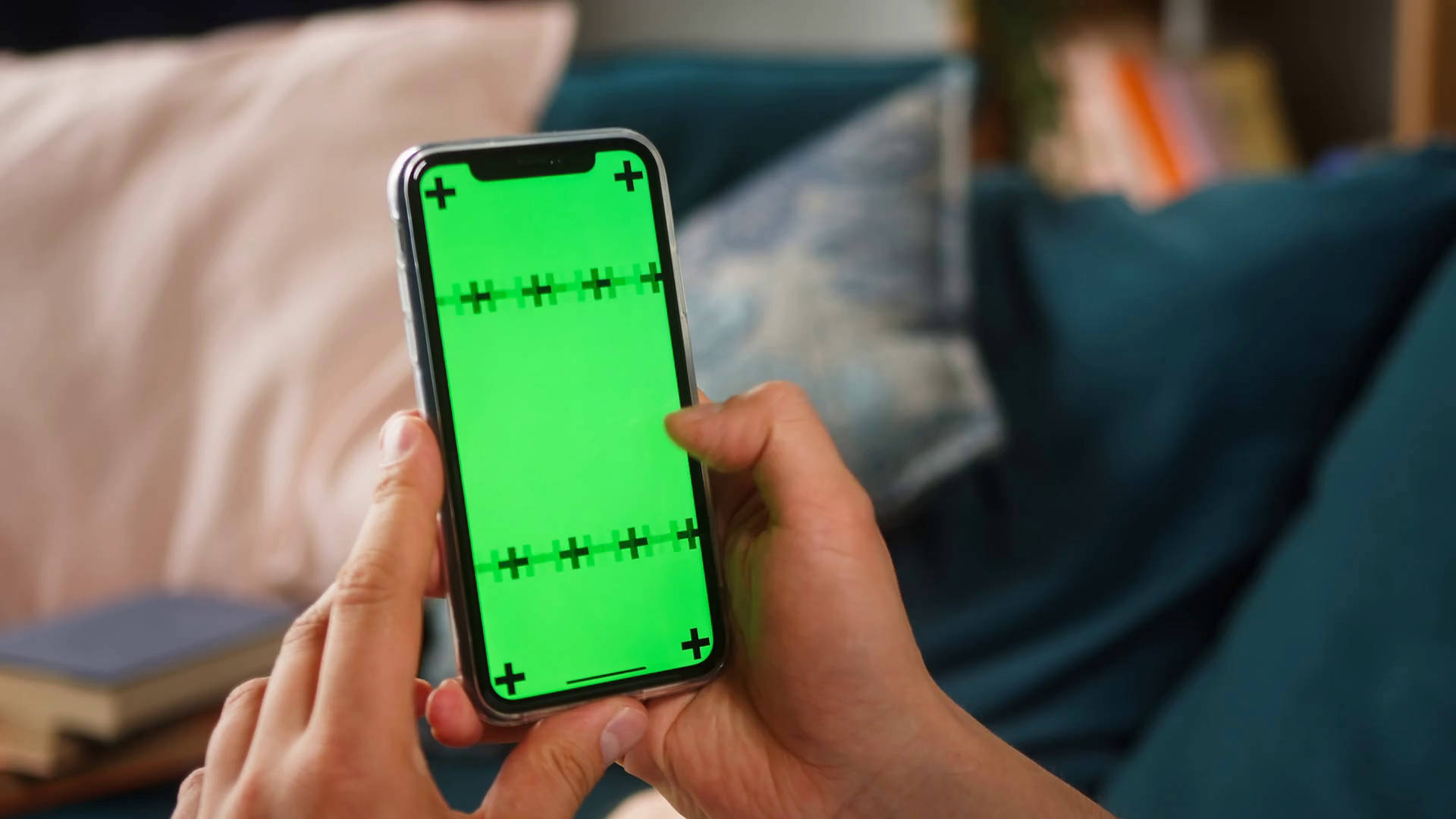 Green Screen 桌布 壁紙 — 雙手握住一支智慧型手機，螢幕顯示綠色，上面有加號和縫合線，背景是模糊的沙發。