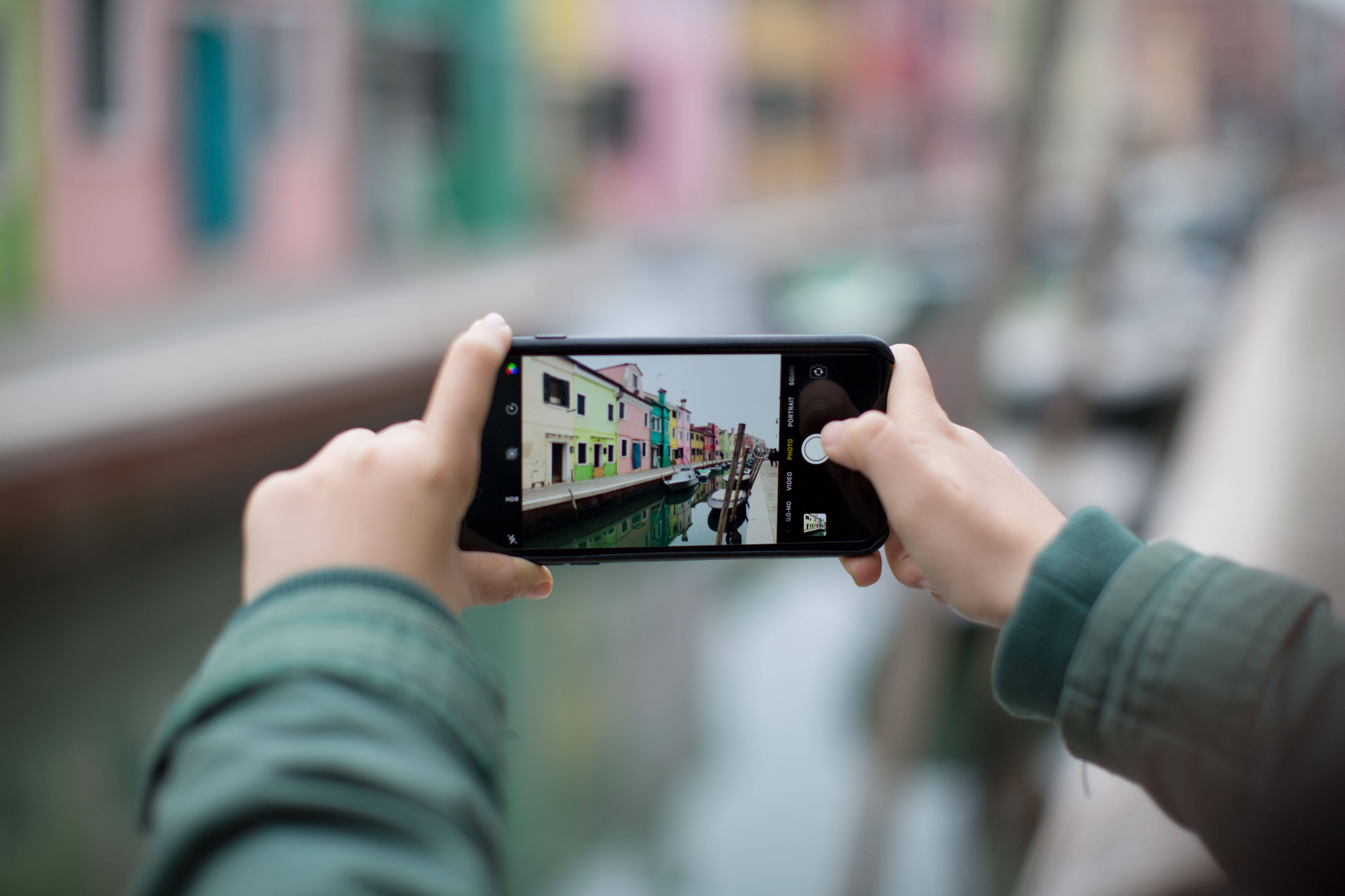 photography iPhone 桌布 壁紙 — 雙手握住智慧型手機，捕捉色彩繽紛的建築物倒映在水中的運河場景。