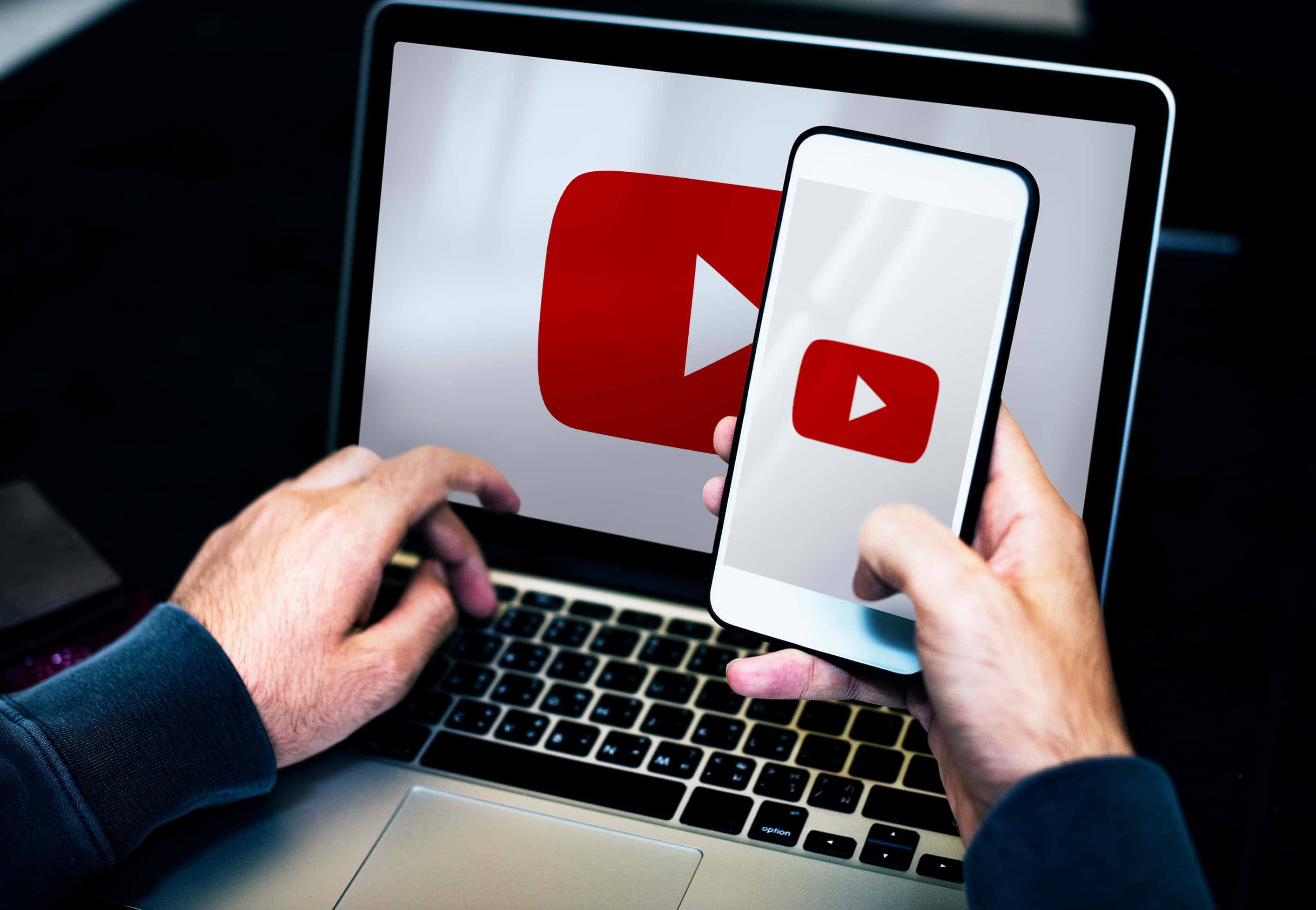 YouTube 桌布 壁紙 — 雙手握住智慧型手機和筆記型電腦，兩者都在昏暗的房間中顯示YouTube標誌。