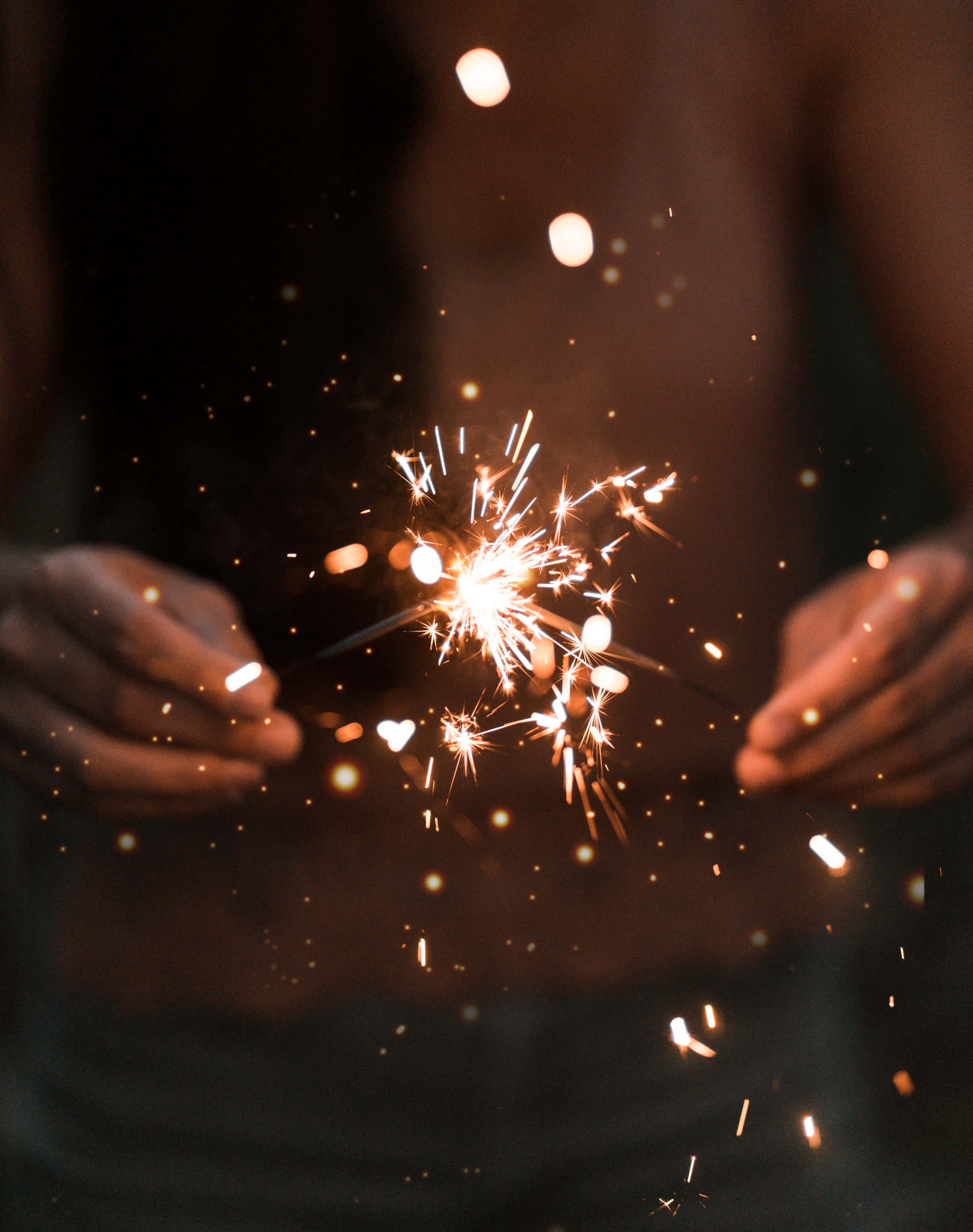 sparklers 桌布 壁紙 — 雙手握著閃爍的煙火，在黑暗模糊的背景中釋放出明亮的金色火花。