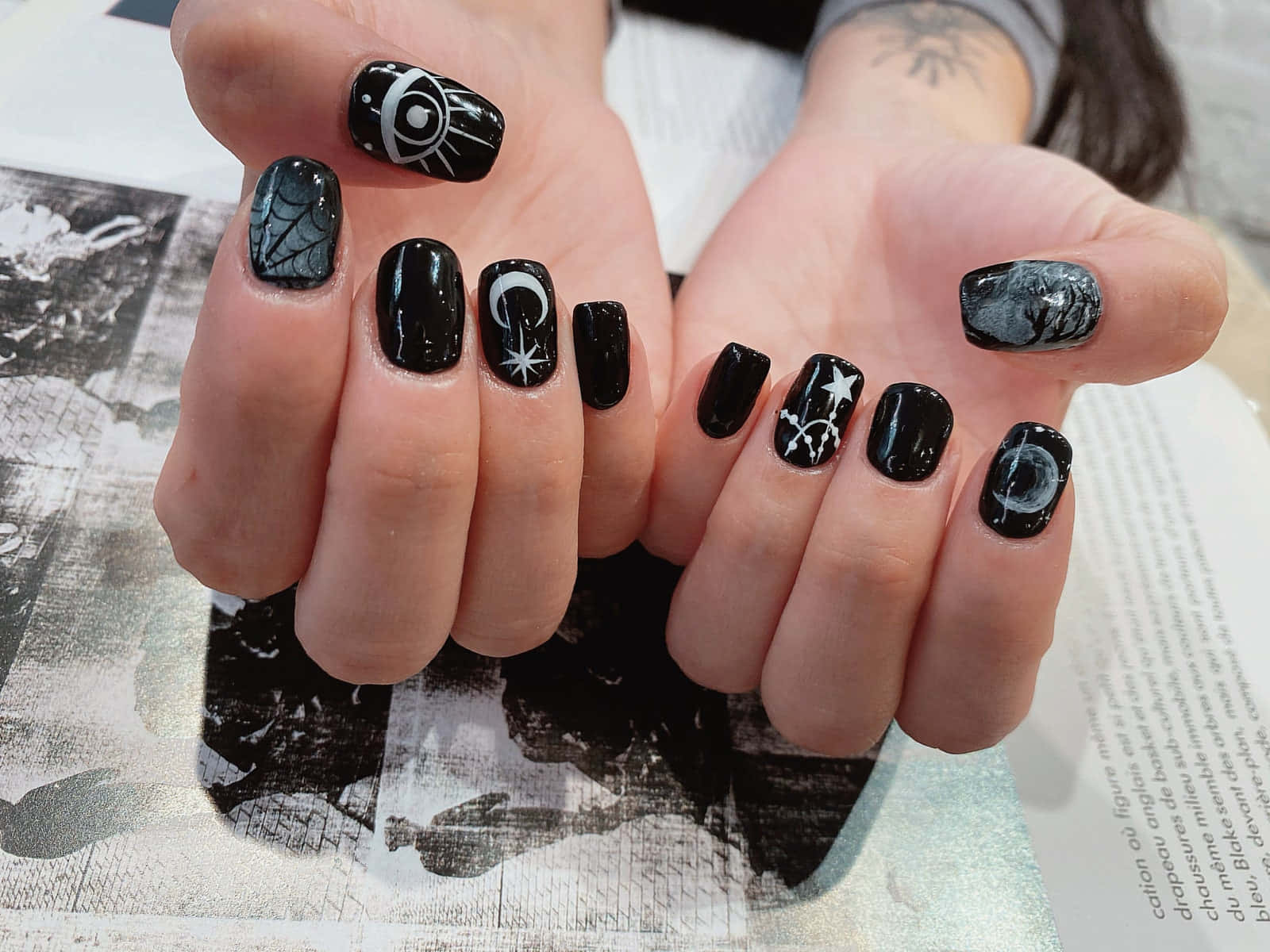 Halloween Nail Art 桌布 壁紙 — 雙手特寫，帶有複雜的黑白神秘指甲藝術，包括眼睛、月亮和星星，背景為有紋理的報紙。