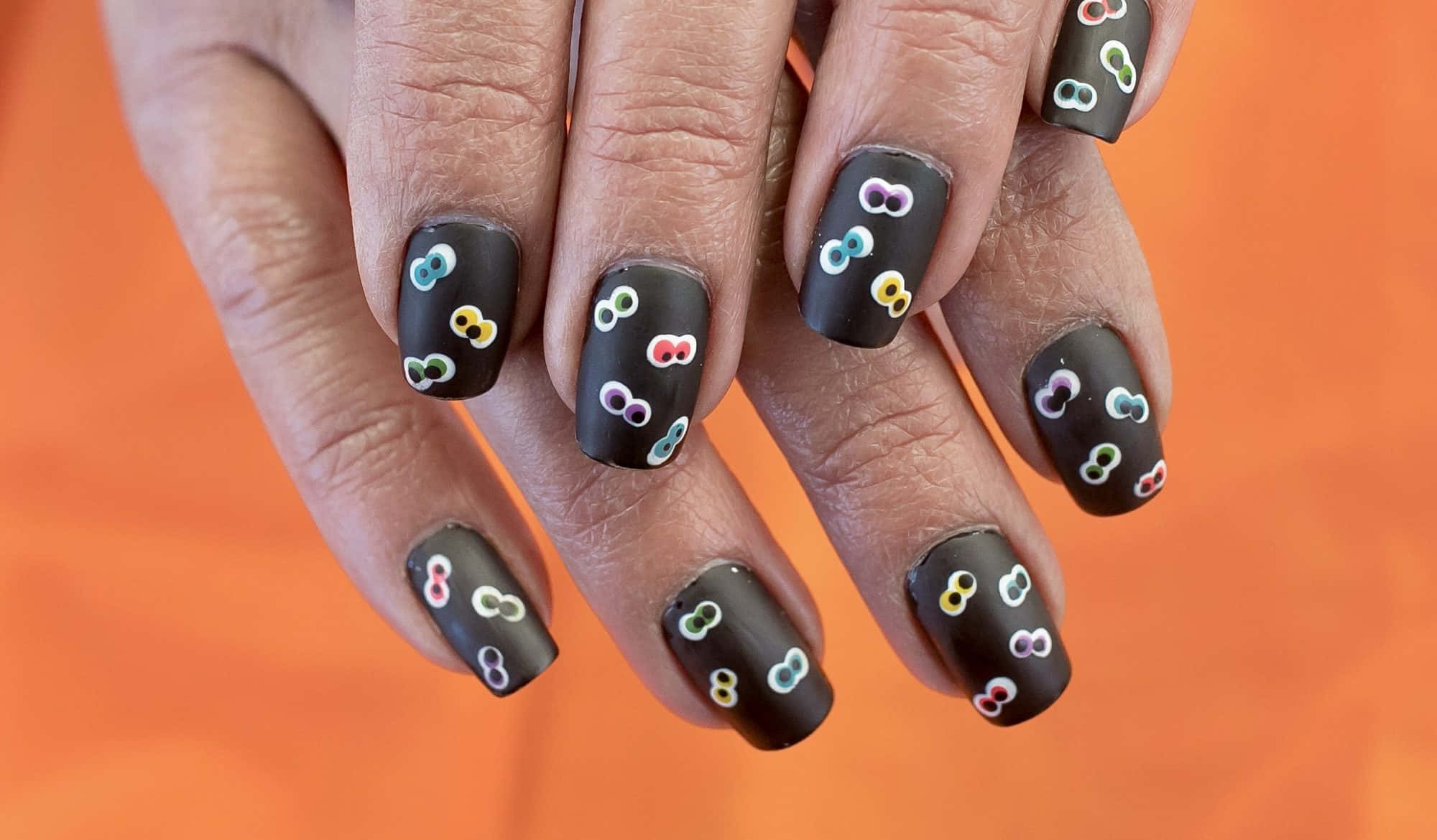 Halloween Nail Art 桌布 壁紙 — 雙手特寫，黑色霧面指甲上飾有色彩繽紛的小無限符號，背景為鮮豔的橙色。