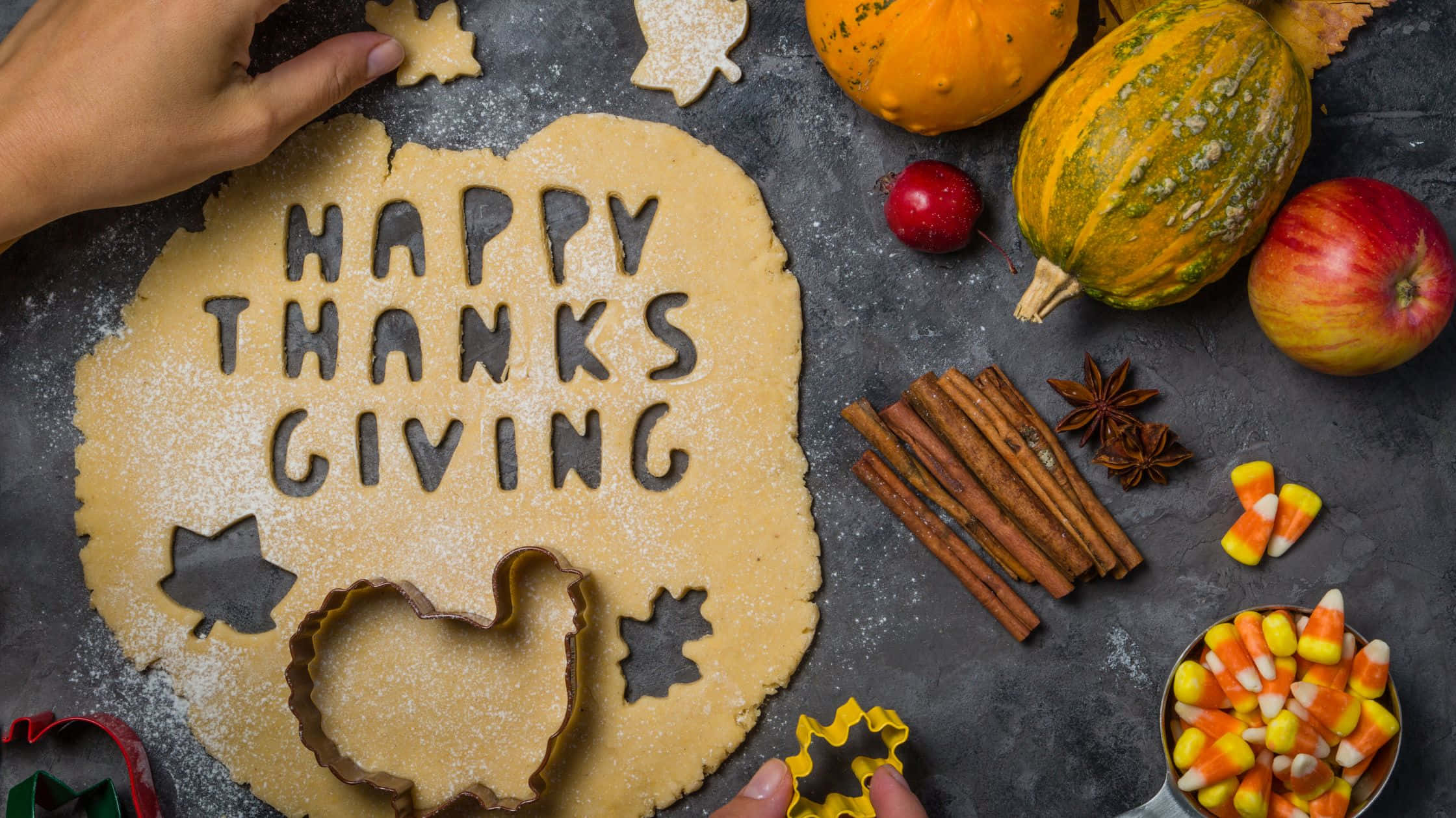 感恩節 桌布 壁紙 — 雙手用秋天主題模具將麵團塑成「HAPPY THANKSGIVING」字樣，周圍環繞南瓜、蘋果和香料。