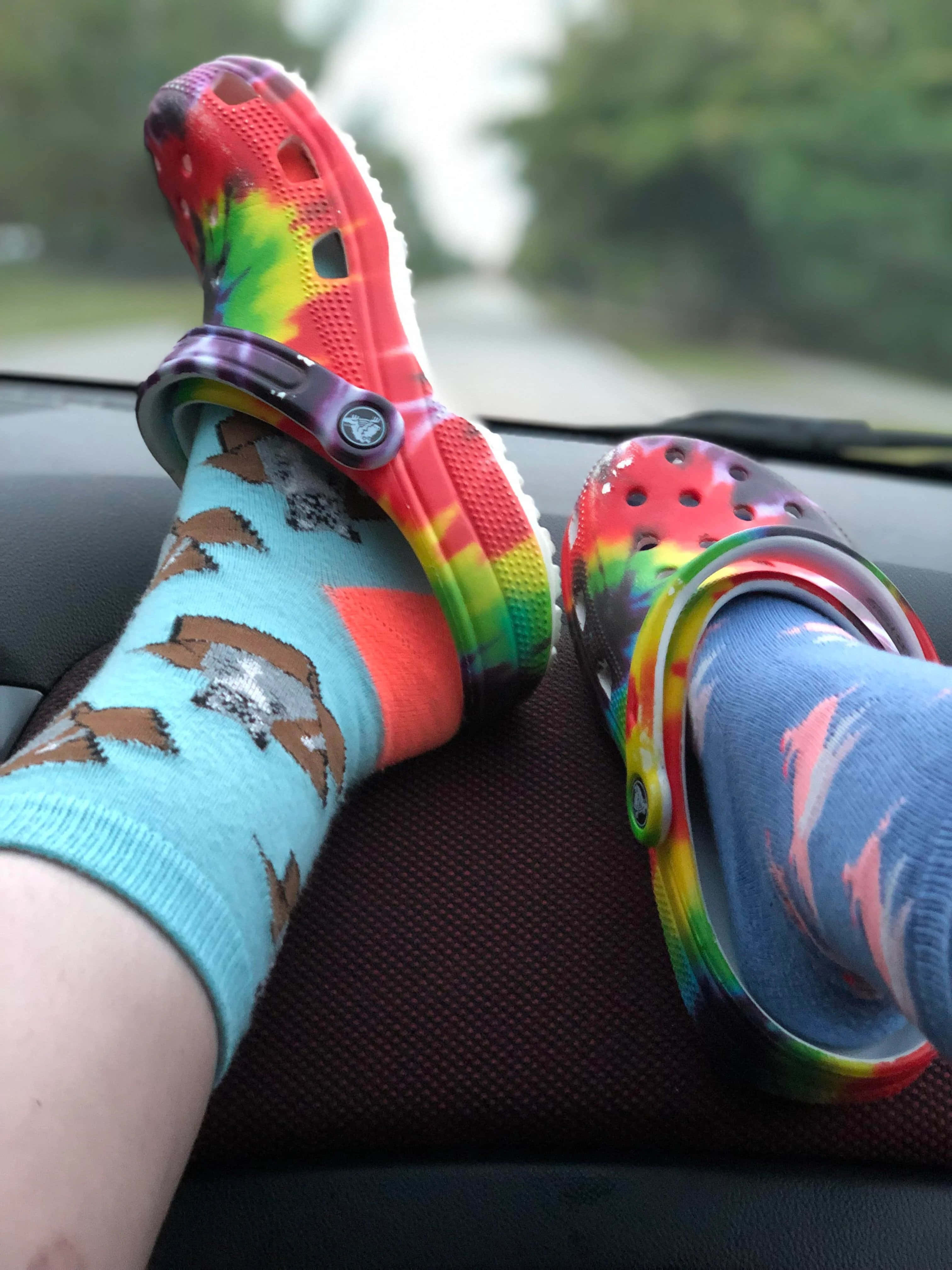 Crocs 桌布 壁紙 — 雙腳穿著彩虹紮染Crocs和圖案襪子，擱在汽車儀表板上，前方是模糊的綠色道路。